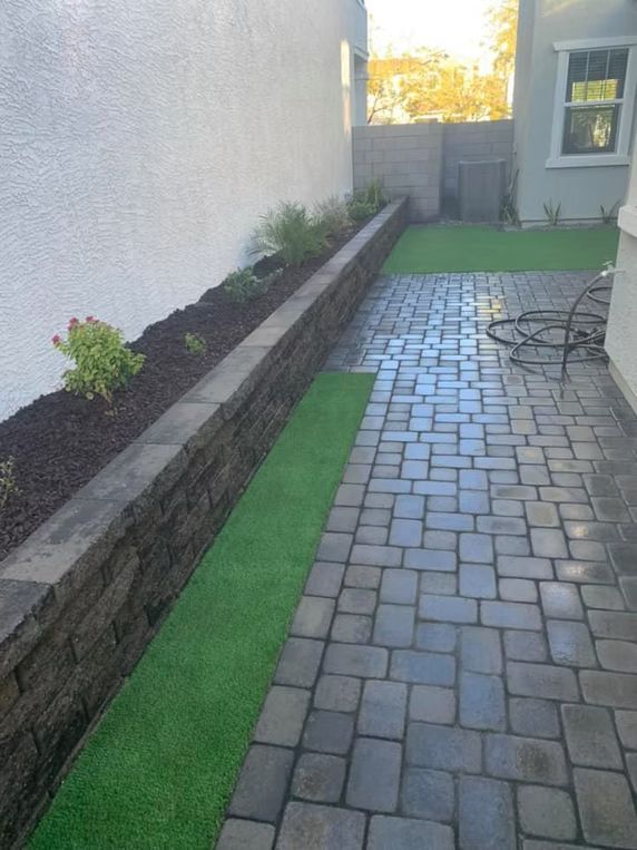 Stone patio next to a narrow strip of green turf and a retaining wall with mulch and plants.