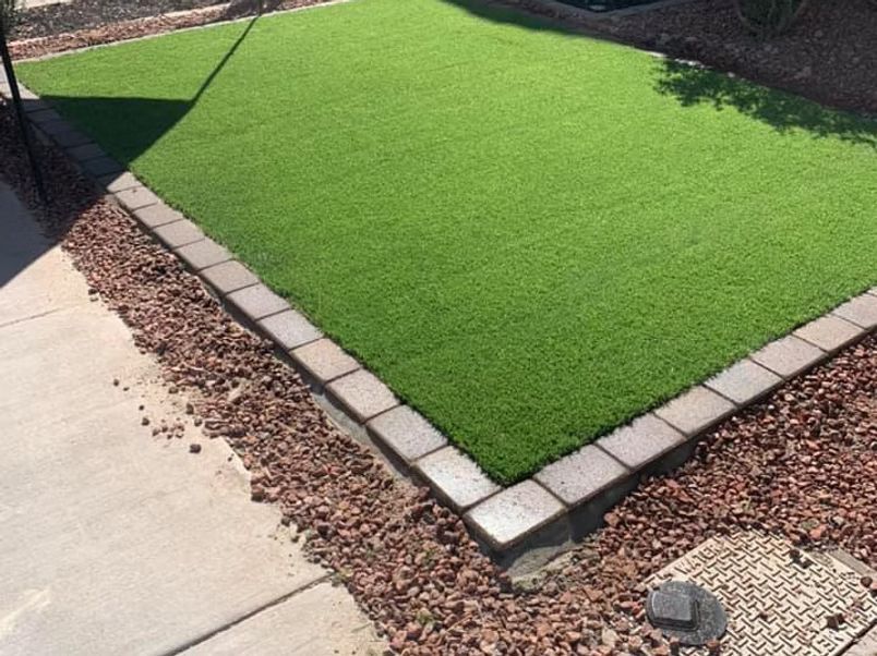 A square patch of bright green artificial turf surrounded by a brick border and red gravel.
