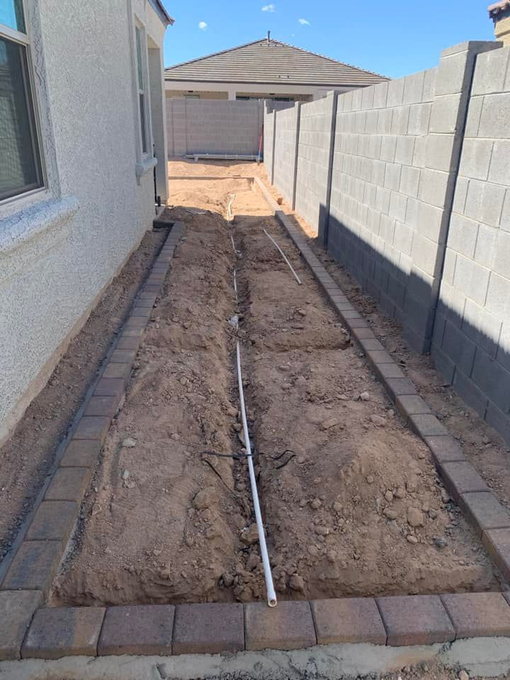 Narrow yard with brick-lined dirt beds, a white pipe, and a cinder block wall under a clear sky.