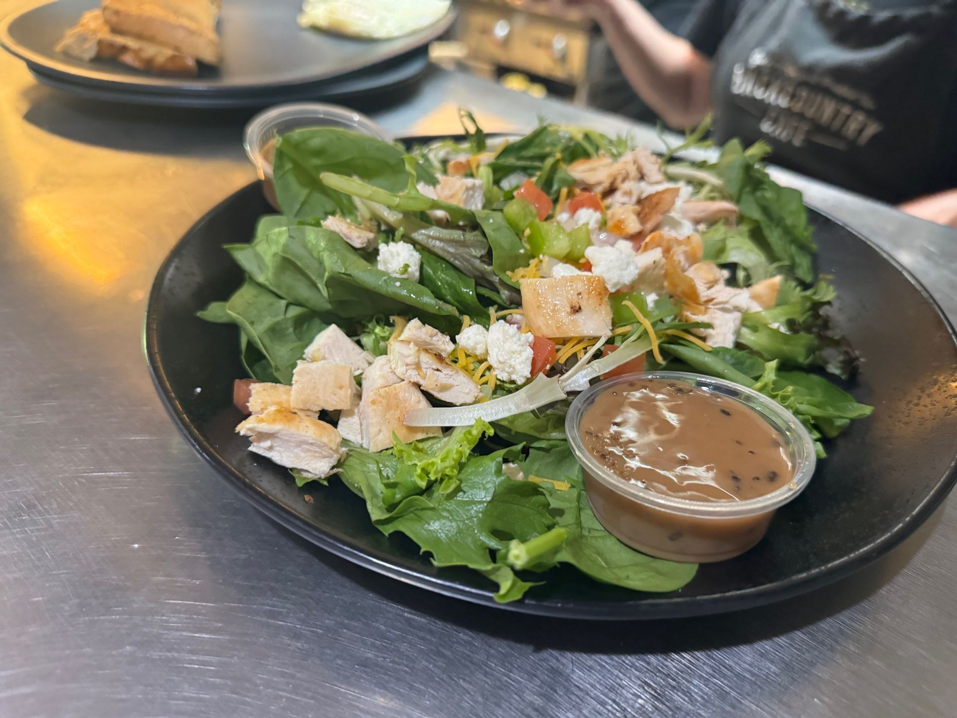 A salad with chicken and spinach on a black plate with a cup of dressing.