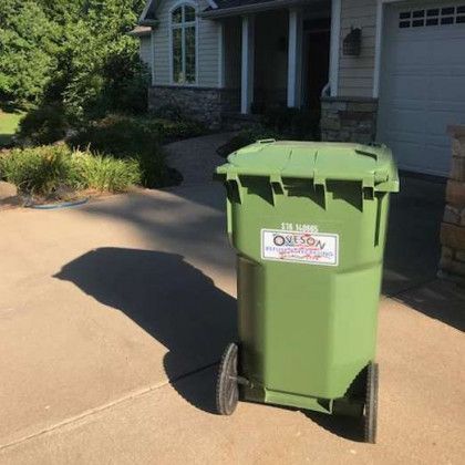 Green trash bin on wheels in a driveway, with a house and greenery in the background.