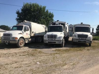 Three white garbage trucks parked on a dirt lot under a clear blue sky.