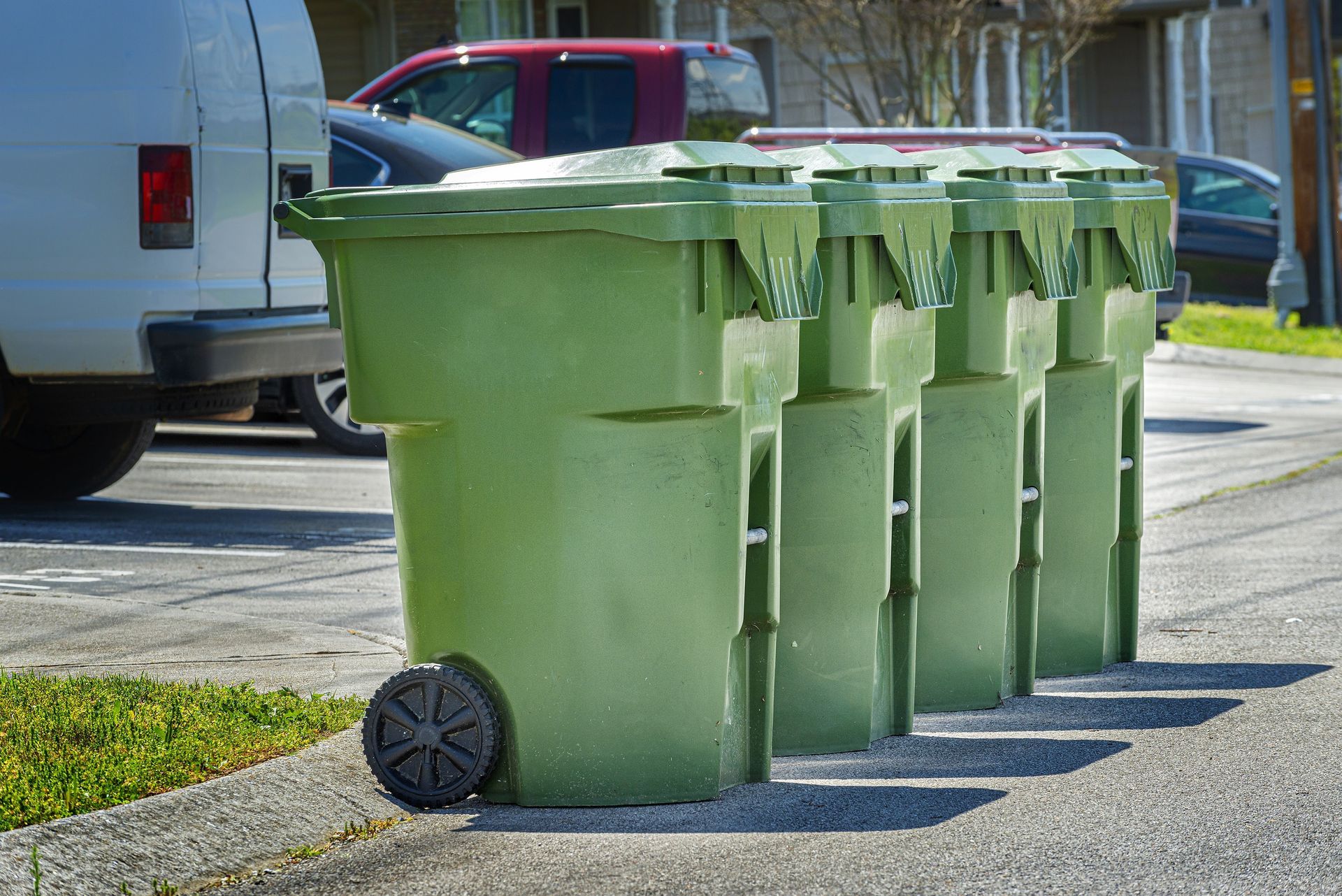 Green trash cans lined up on a sidewalk with vehicles in the background.
