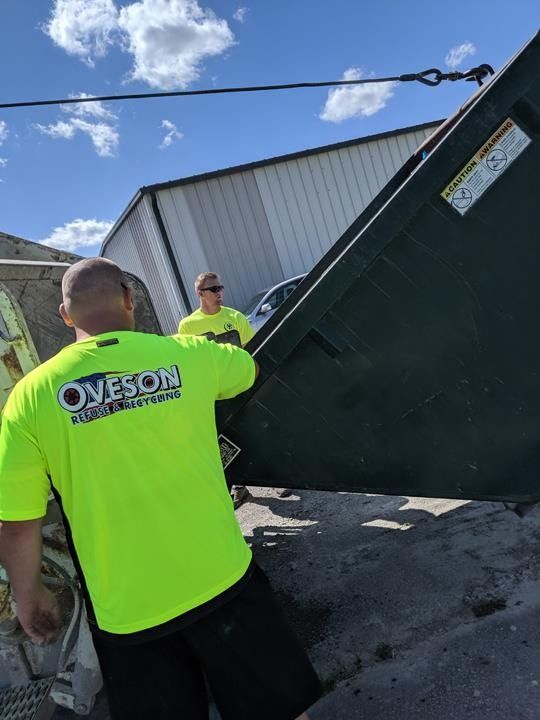 Two men in neon shirts emptying a dumpster under a blue sky.