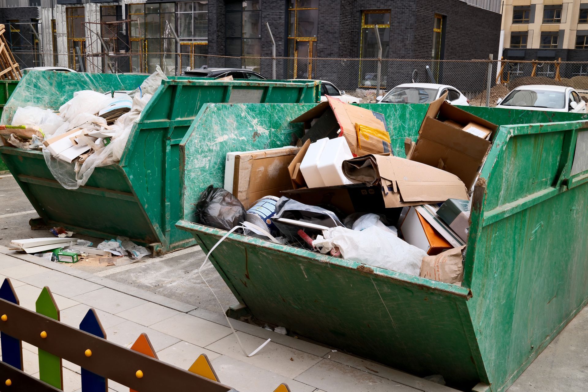 Two overflowing green dumpsters in a parking area, filled with trash and cardboard.