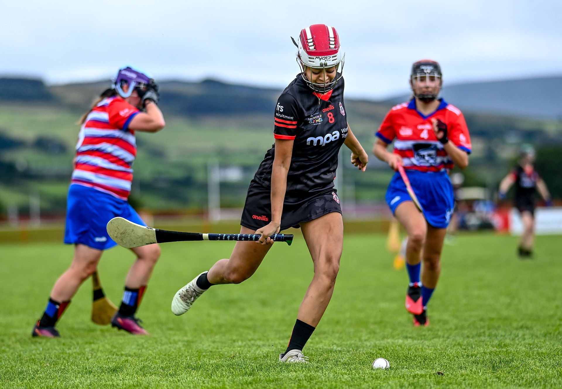 Female camogie players in action on a green field. Players in helmets and uniforms, holding hurleys and chasing a ball.
