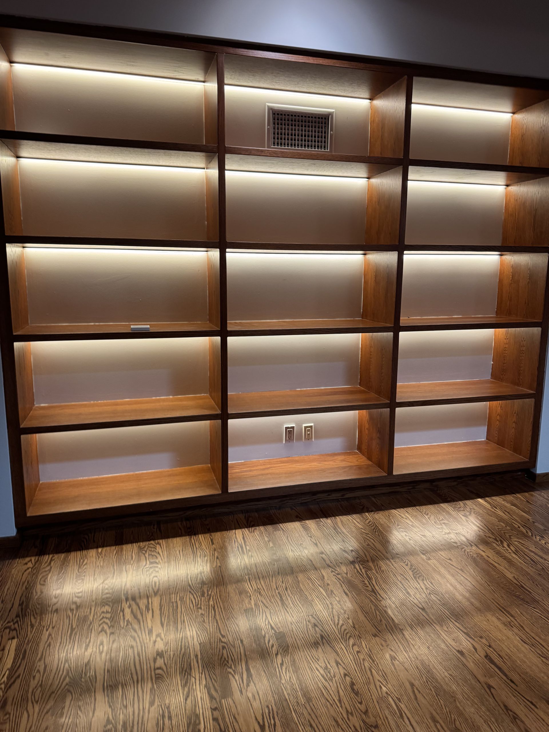 Wooden built-in bookshelf with LED lighting illuminating shelves, on a hardwood floor.
