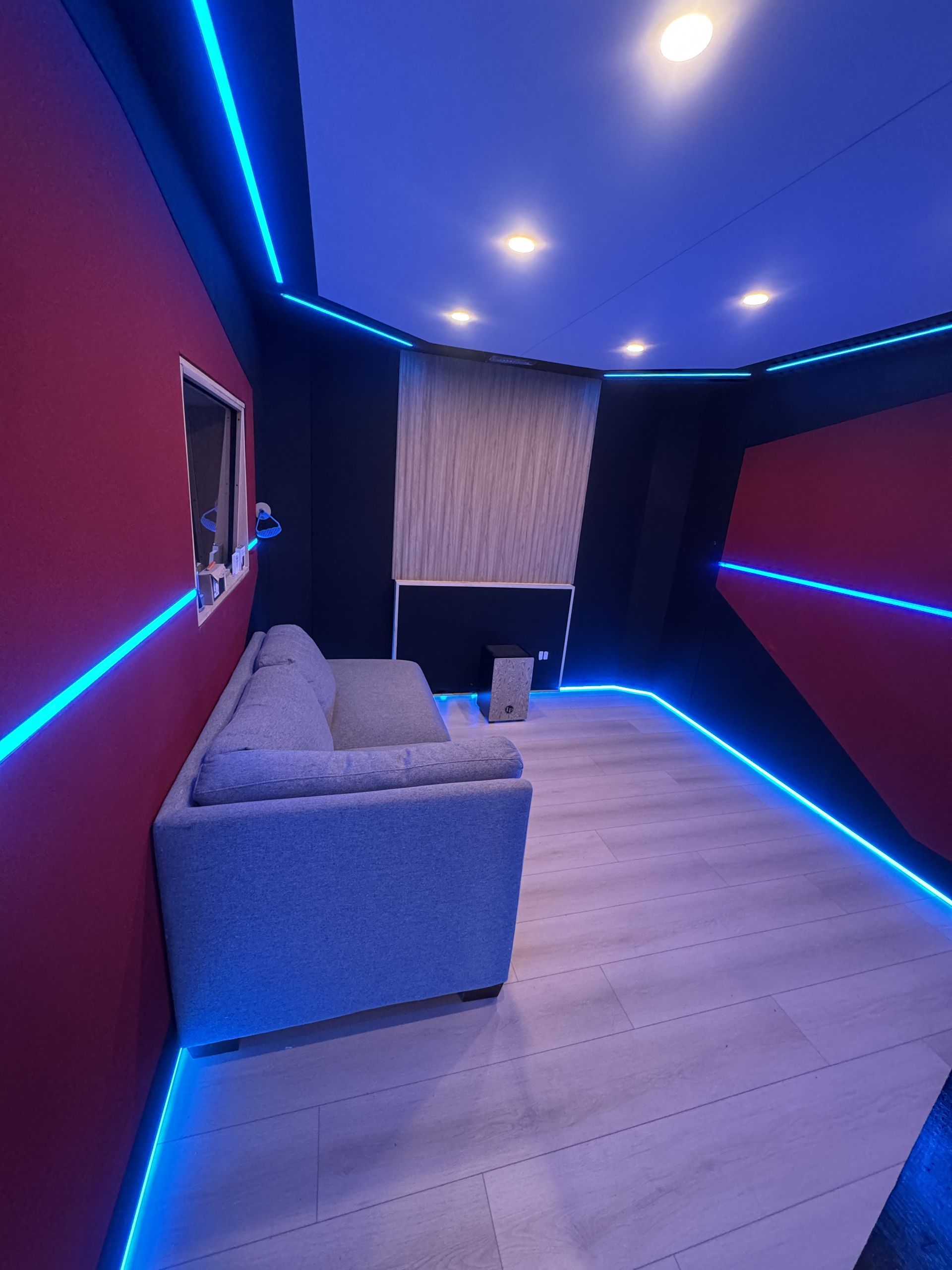 Cozy room with gray couch, red and black walls, blue LED lights, and a window with a textured blind.