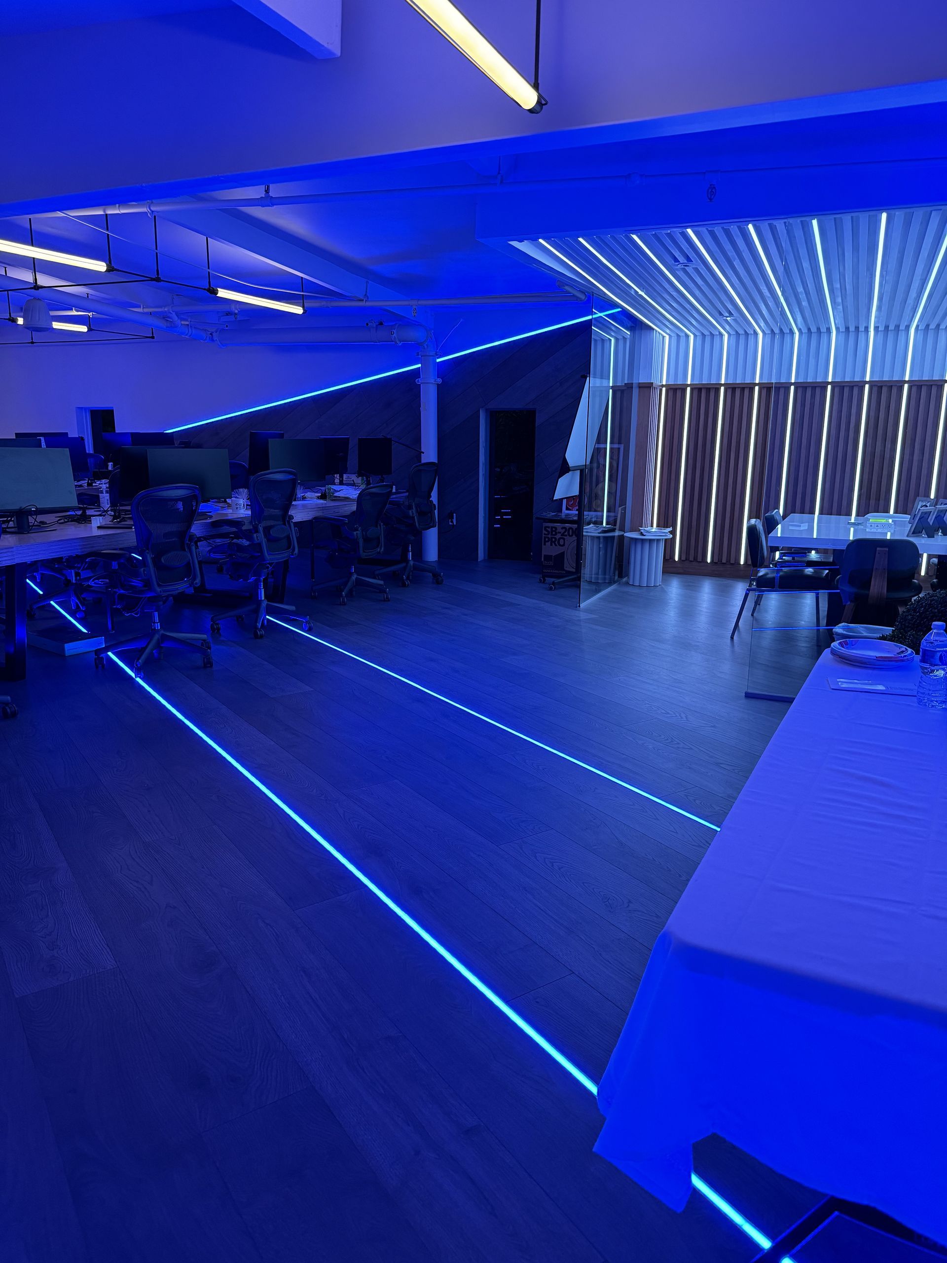 Blue-lit event space with tables, chairs, and linear lighting across ceiling and floor. Wooden panel wall in background.