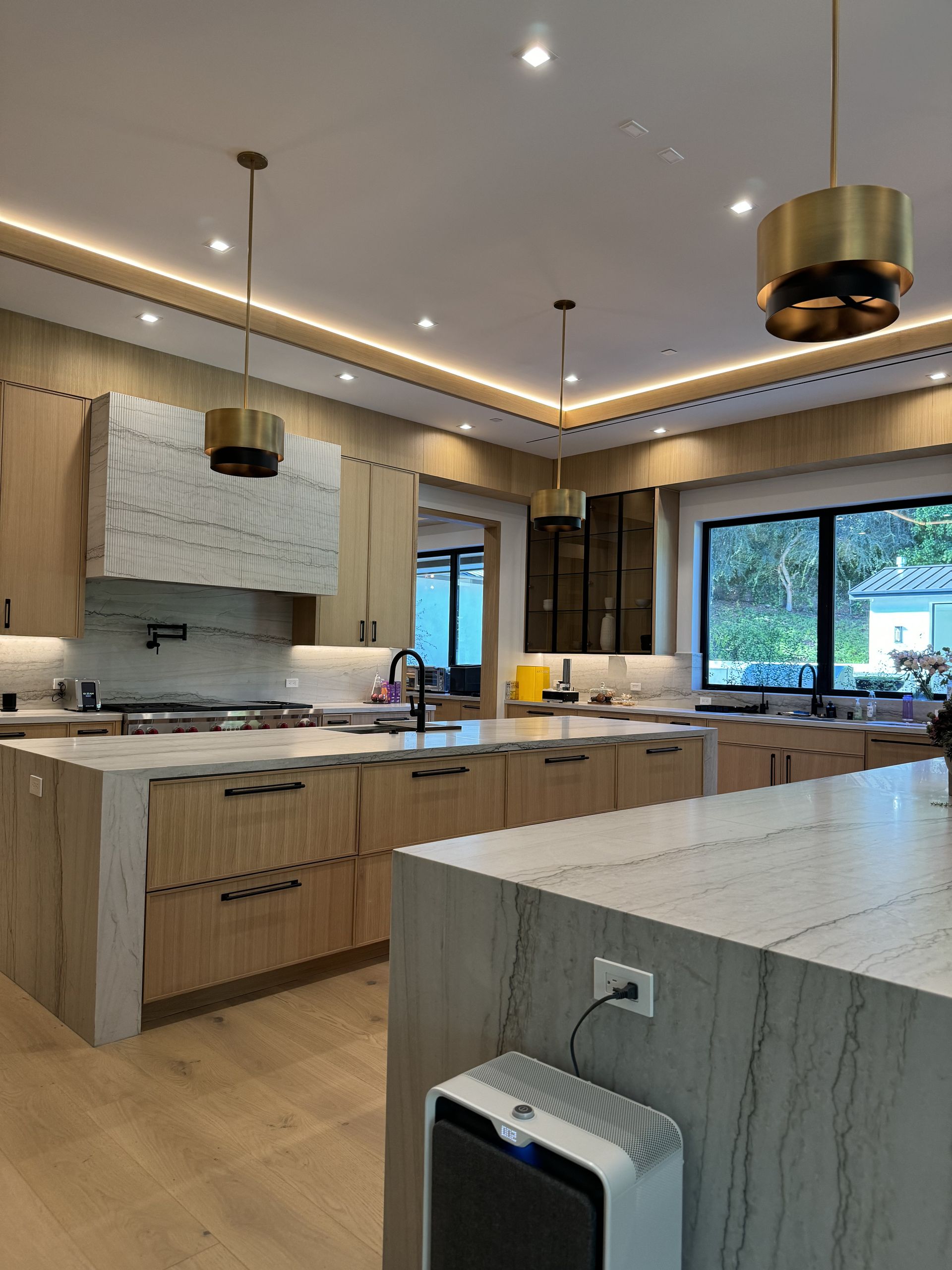 Modern kitchen with light wood cabinets, marble countertops, and gold pendant lights.