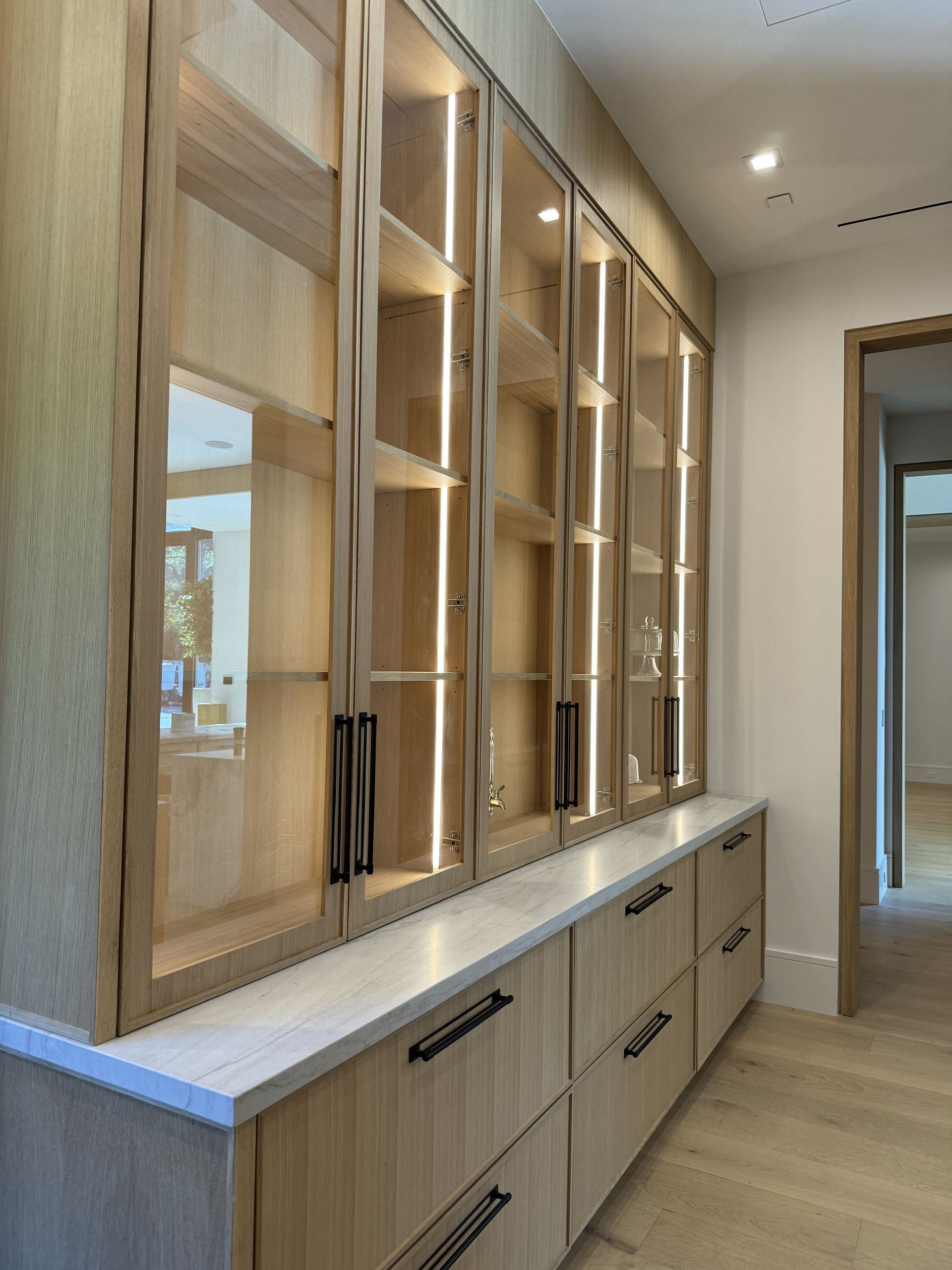 Built-in wooden display cabinets with glass doors, illuminated shelves, and drawers, in a hallway.