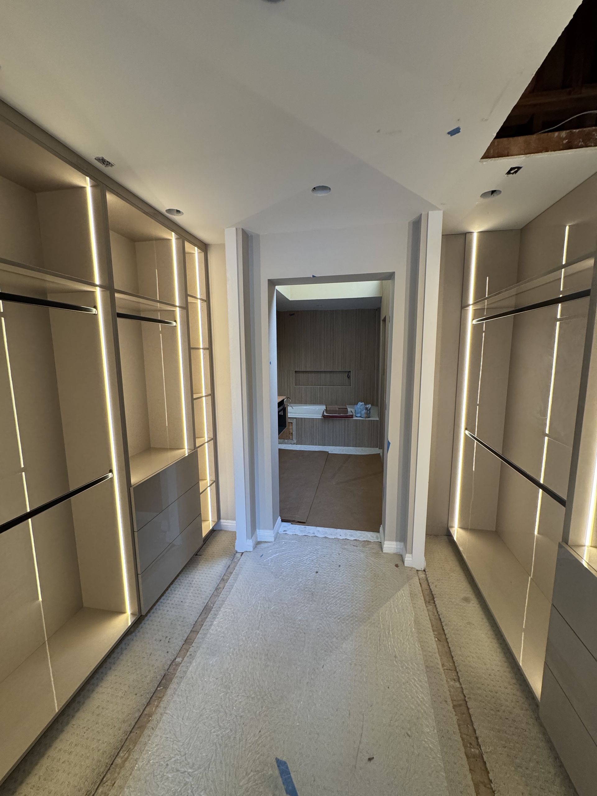 Walk-in closet under construction with recessed lighting. Beige shelving on both sides with doorway at the end.