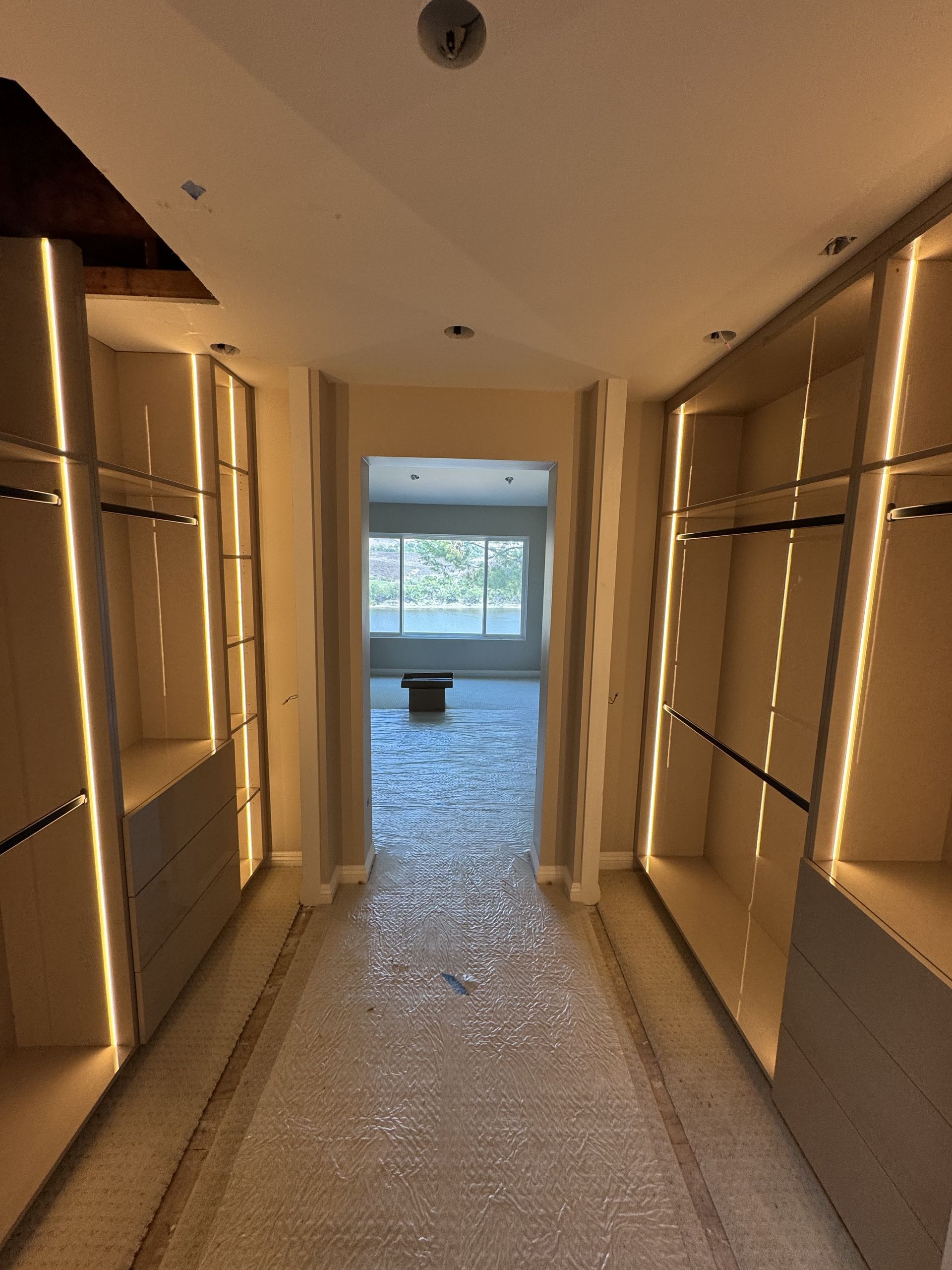 Walk-in closet with beige cabinetry and vertical LED lighting. Open doorway leads to a bright room.