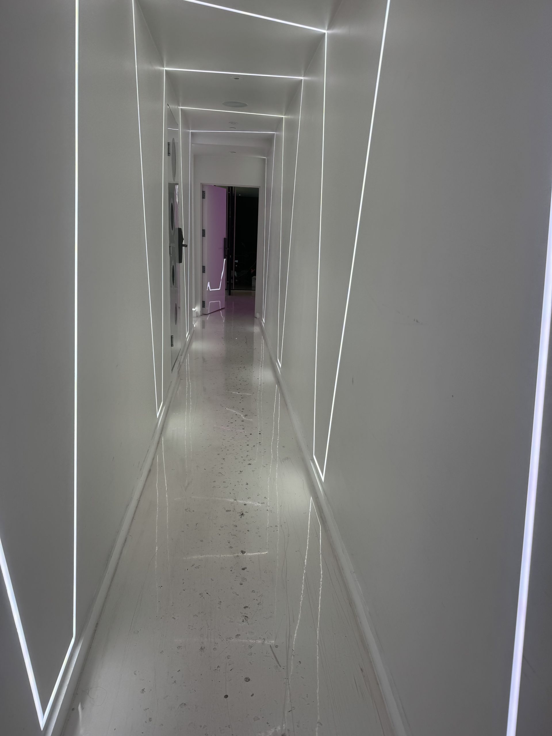White hallway with overhead and side lighting creating patterns and leading to a dark doorway.