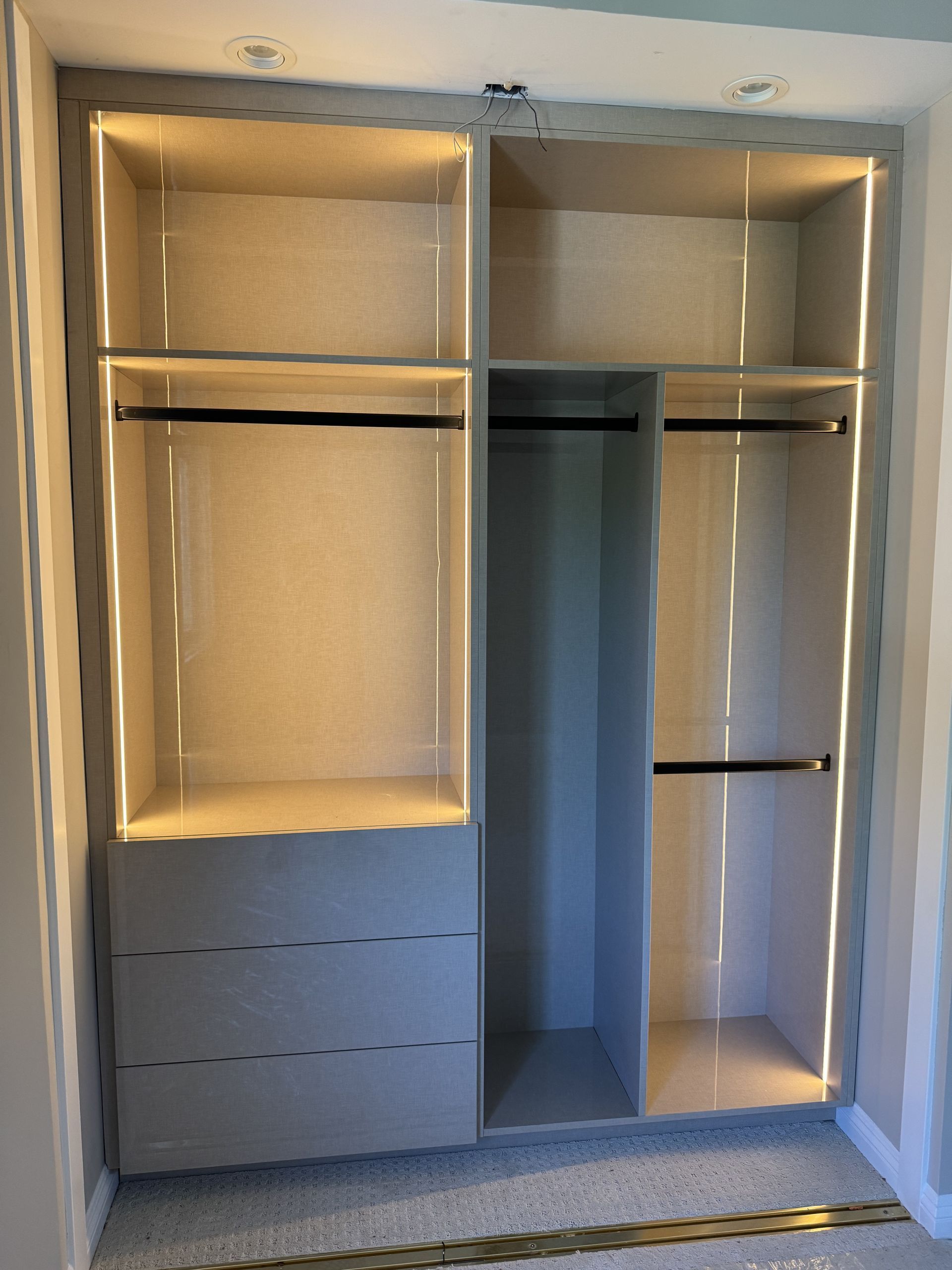 Built-in closet with shelves, drawers, and hanging rods. Lit with warm white LED lights. Light grey finish.
