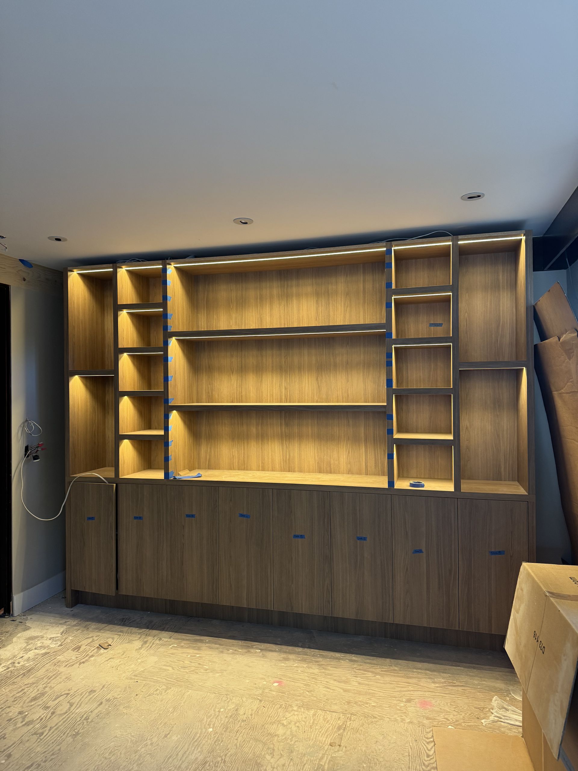 Custom wooden built-in shelving unit with cabinets below and lit shelves. In a room under construction.