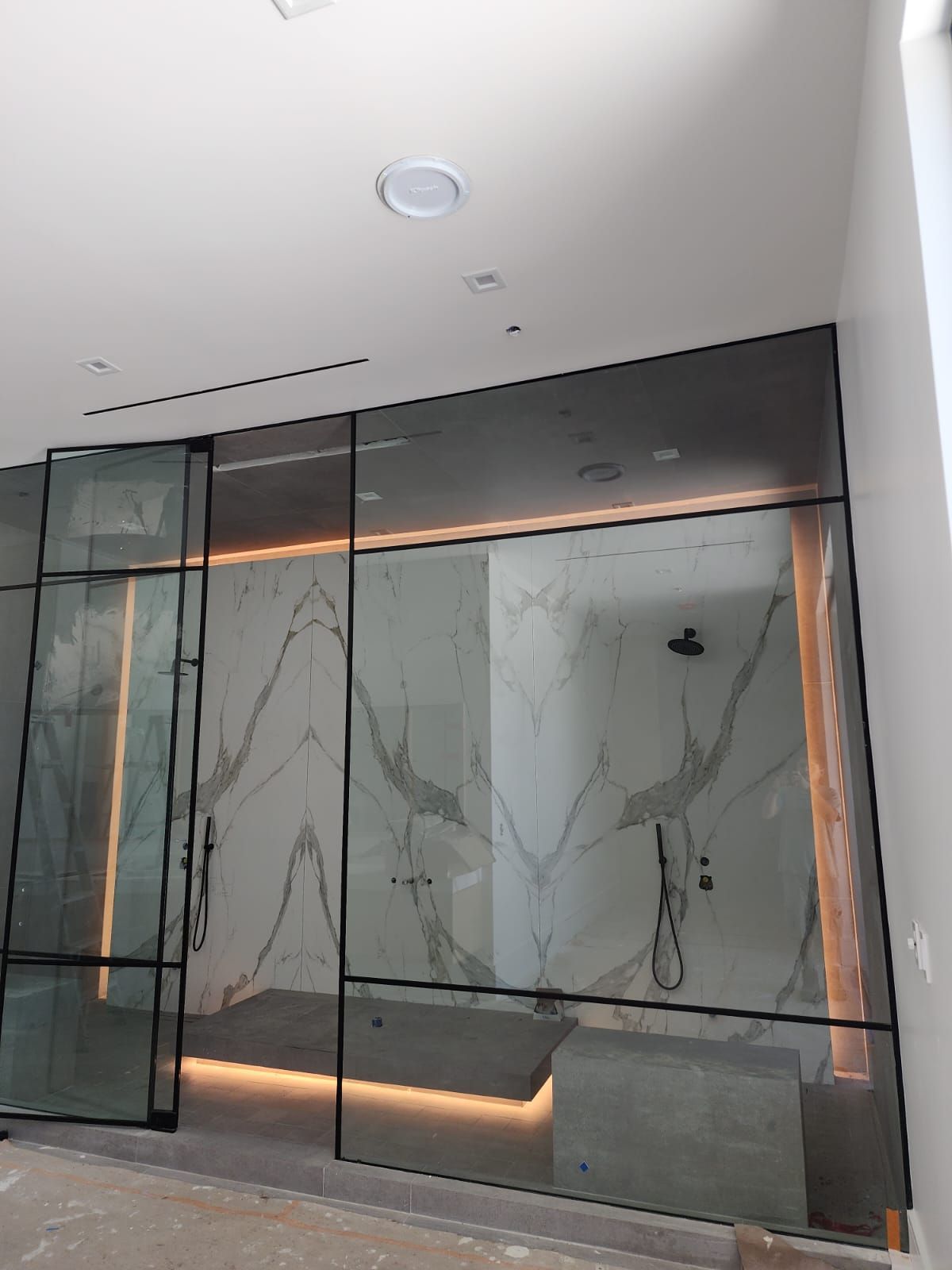 Modern bathroom with glass shower enclosure, marble wall, and linear lighting.