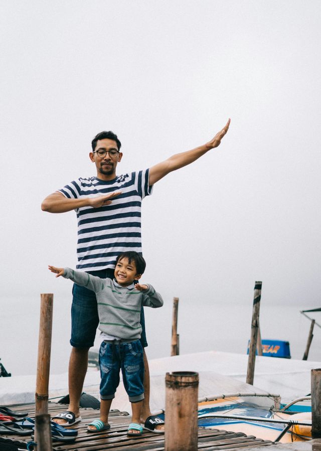 Man and child on a wooden pier, dabbing, cloudy sky. Man wears striped shirt and shorts. Child in gray top and jeans.