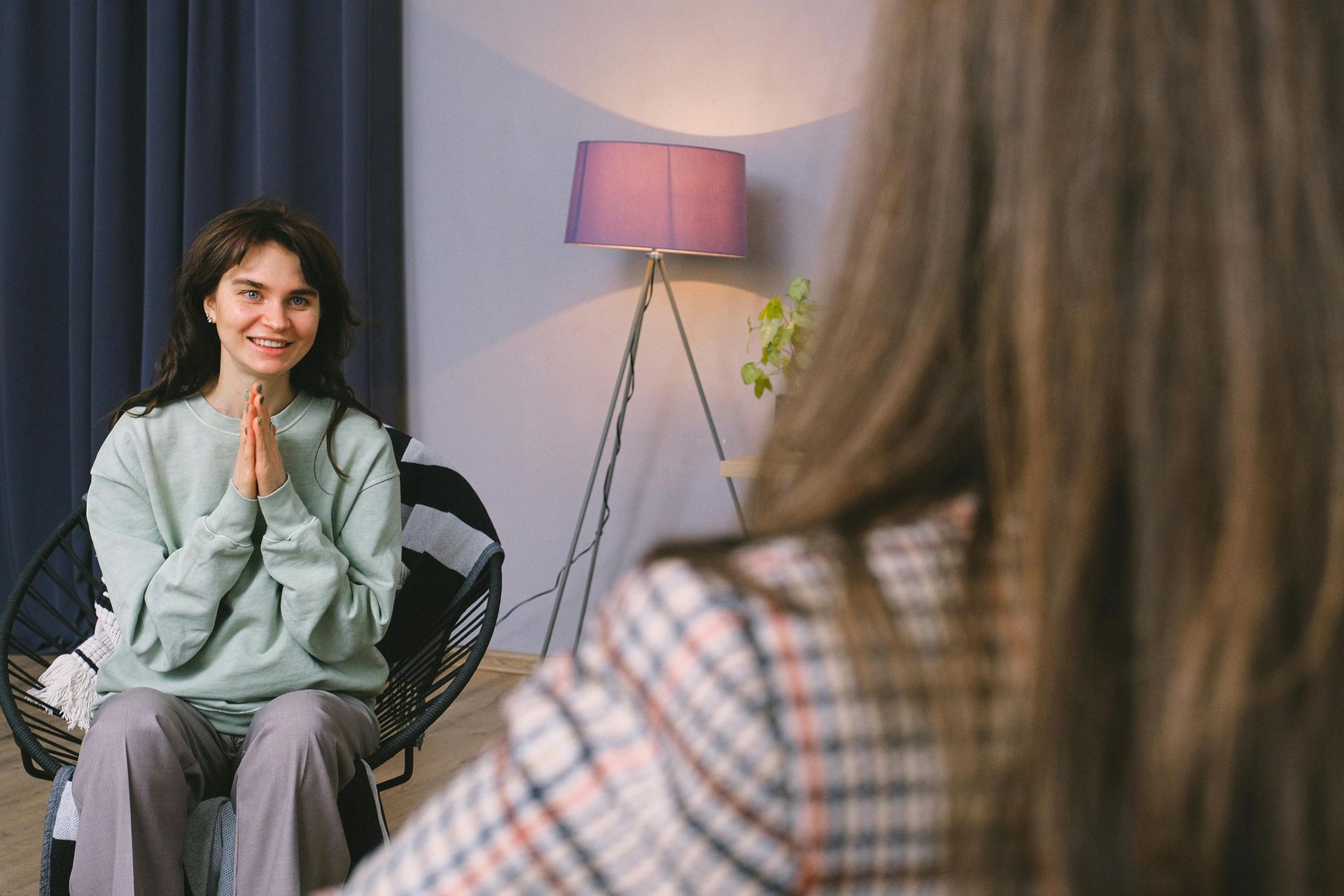 Woman in light green sweatshirt and gray pants smiles, hands clasped, facing a person in a plaid jacket. Indoor setting with lamp.