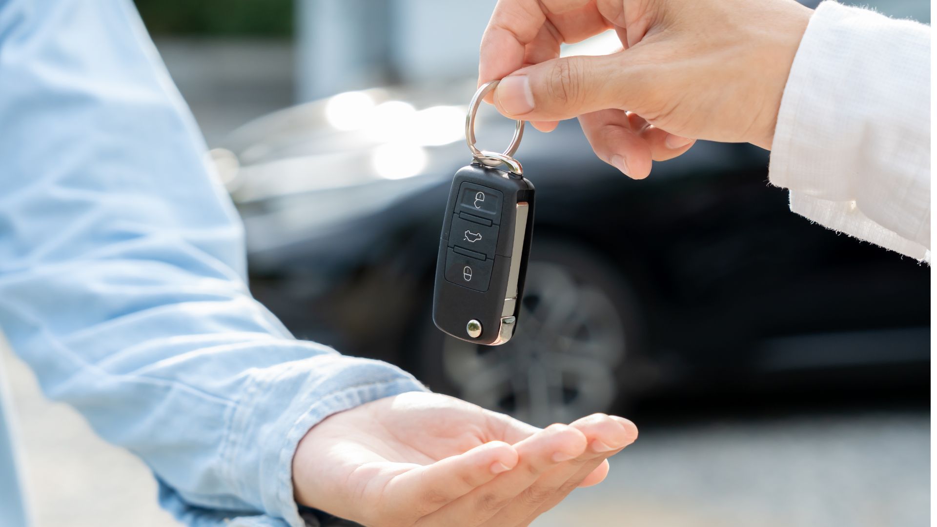 Handing over a car key fob between two people outdoors, with a blurred car in the background