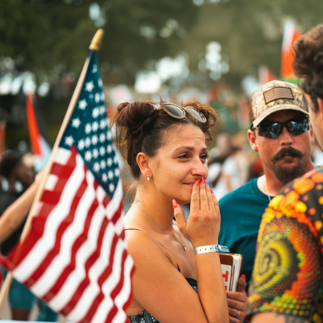 Woman with hand to face, possibly crying, stands near American flag at outdoor event. People in the background.