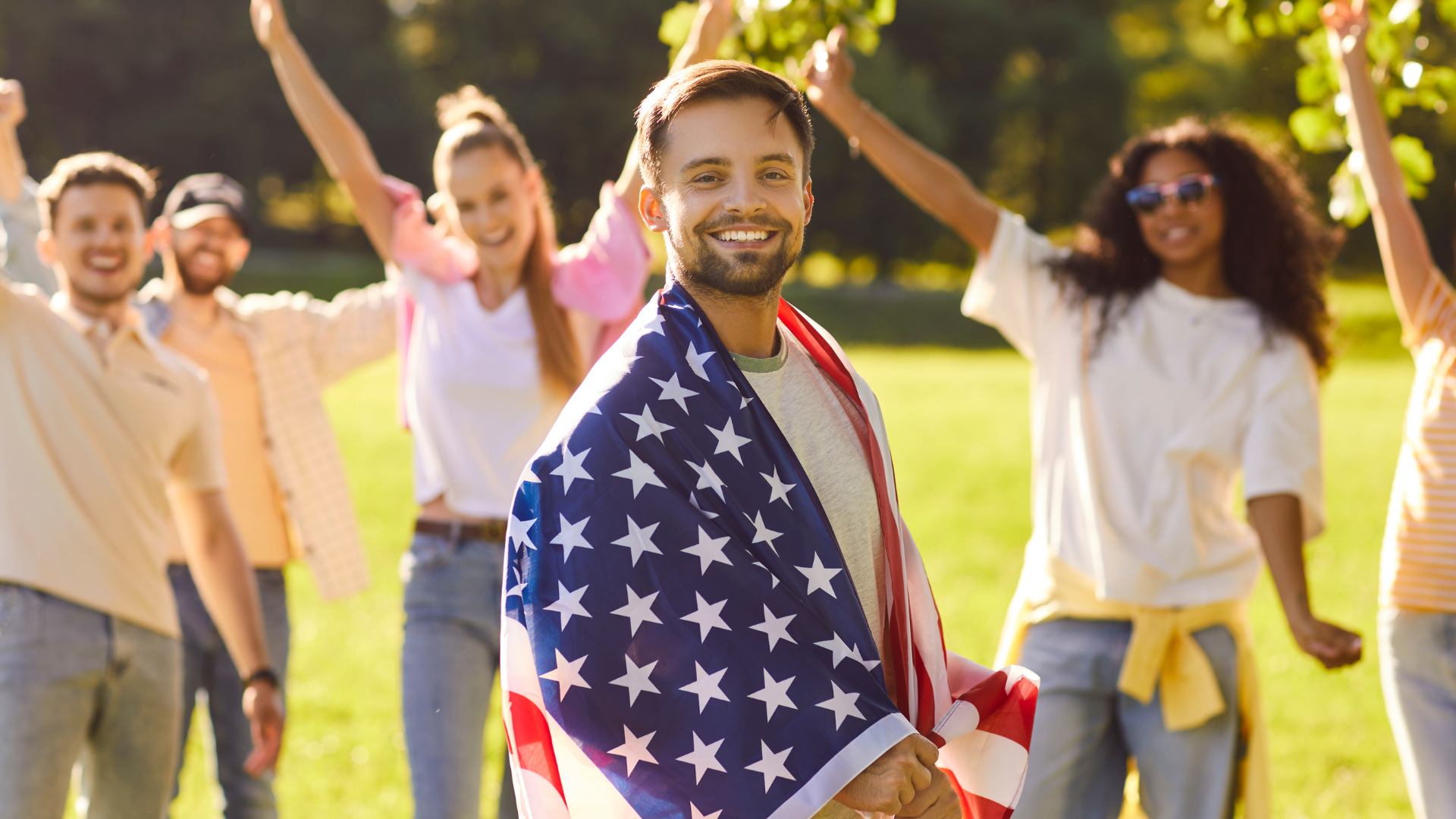 Man smiling, draped in American flag, with group celebrating outdoors.