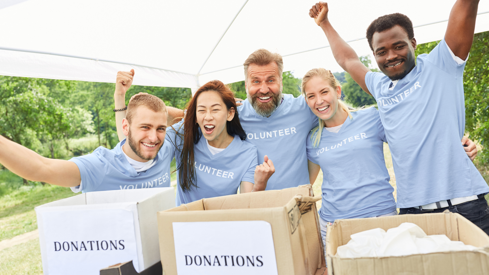 Volunteers cheering, posing behind donation boxes under a white tent; outdoors setting.