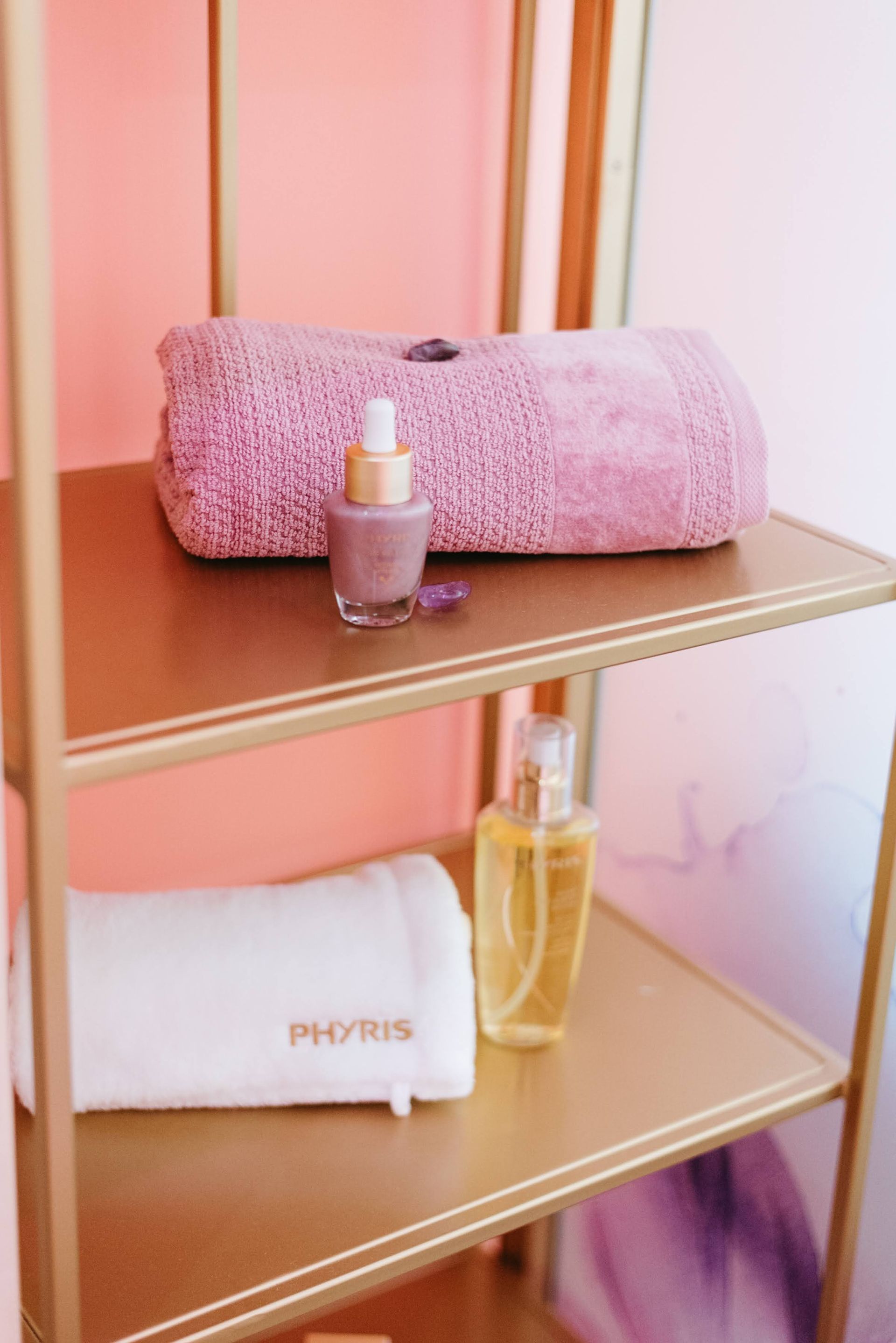 Mensola dorata con prodotti per la cura della pelle e asciugamani nelle tonalità del rosa e del viola.