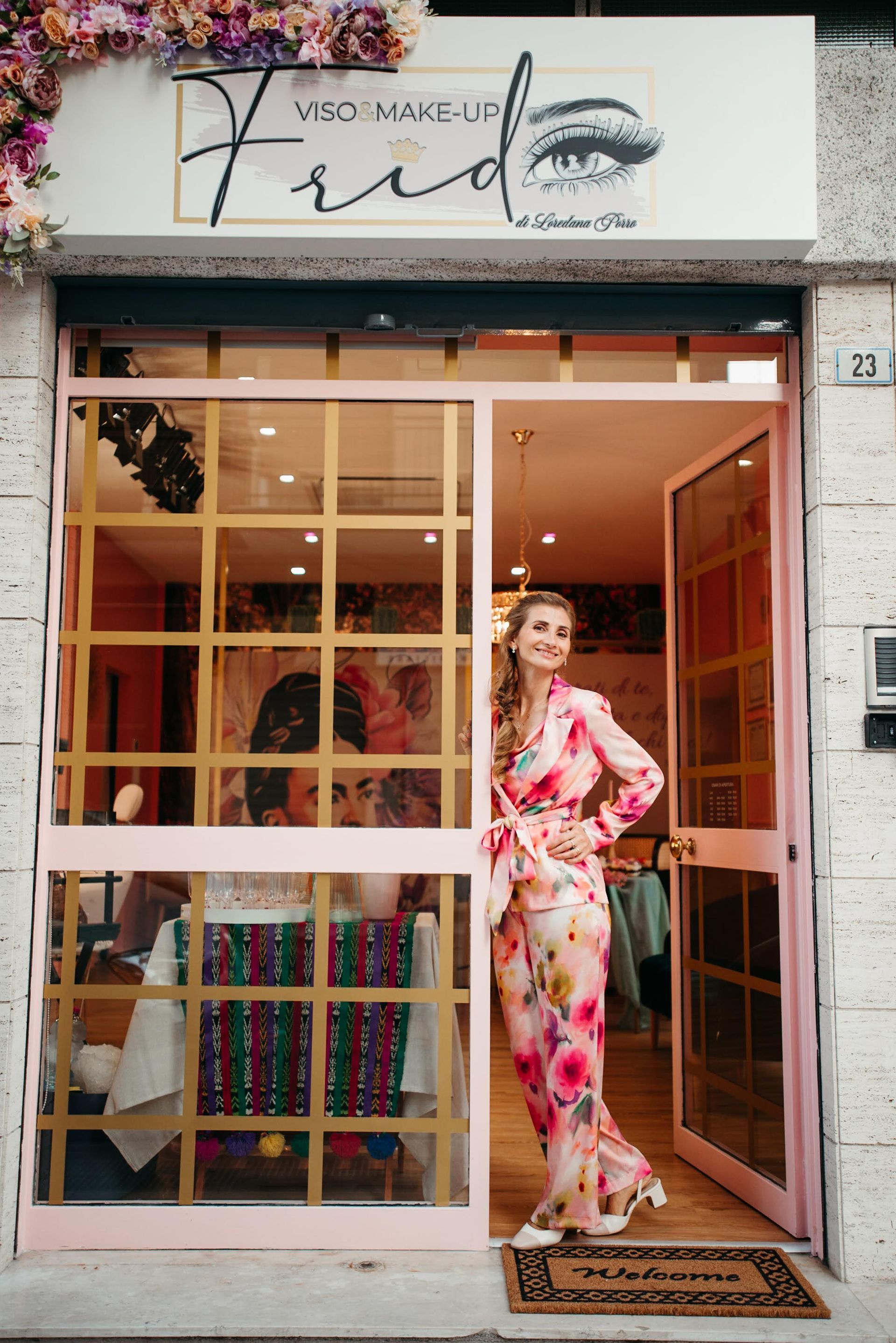 Una donna vestita con un abito floreale è in piedi sulla soglia di un negozio con cornice rosa; l'insegna sulla vetrina recita 