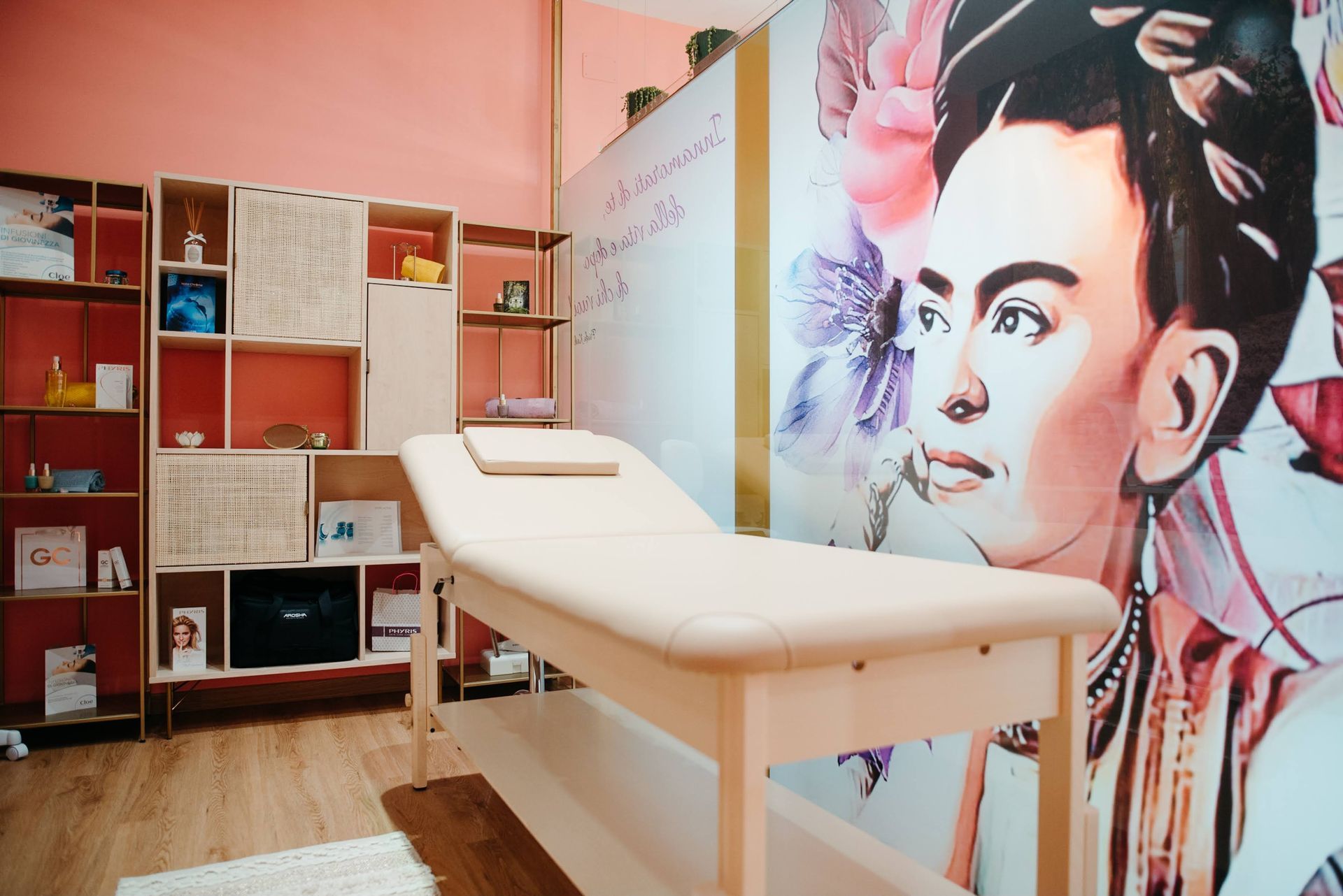 Sala trattamenti con murale di Frida Kahlo, lettino per massaggi beige, scaffali e pareti rosa.