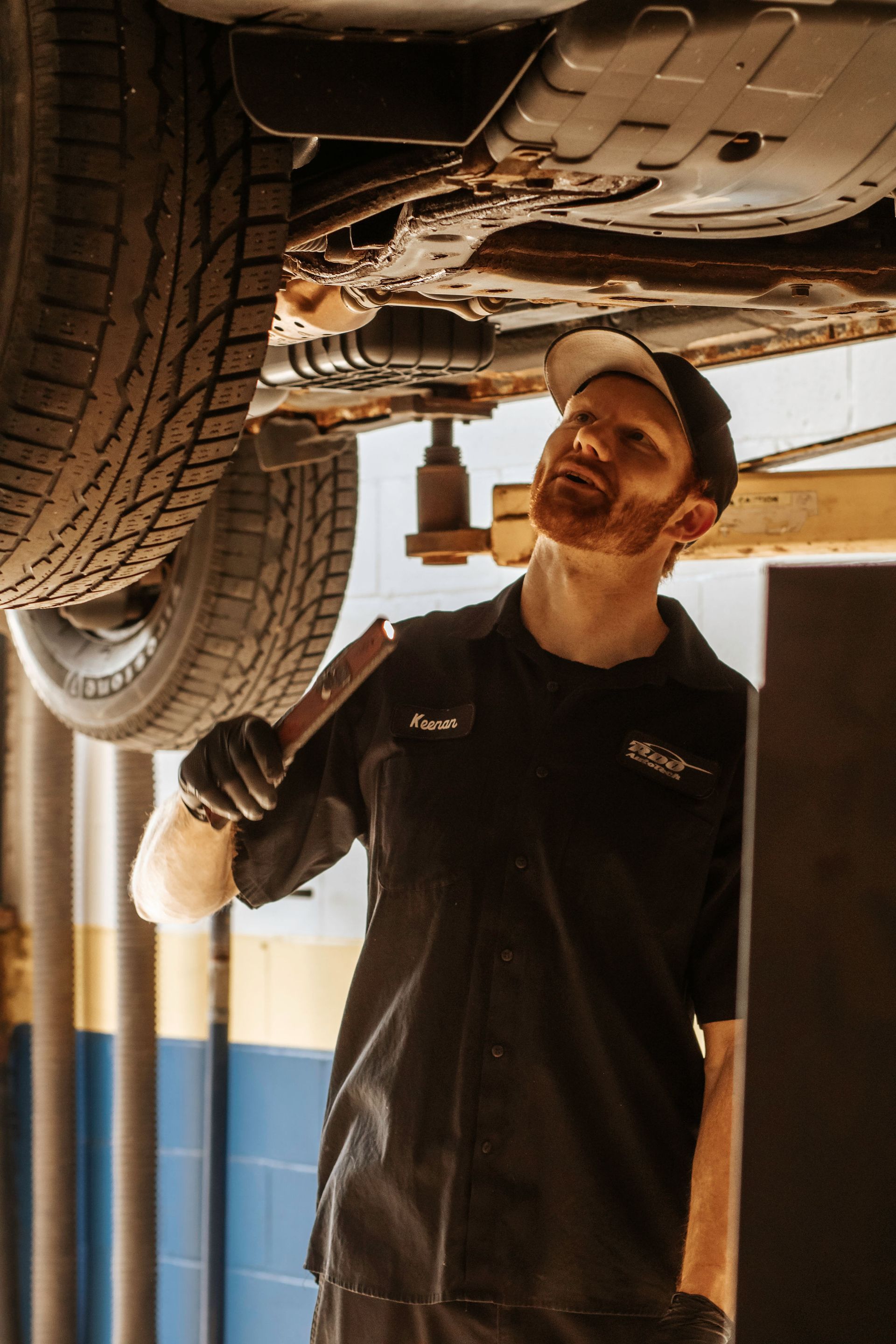 Mechanic in black shirt examines car's underside in auto shop, holding a tool.