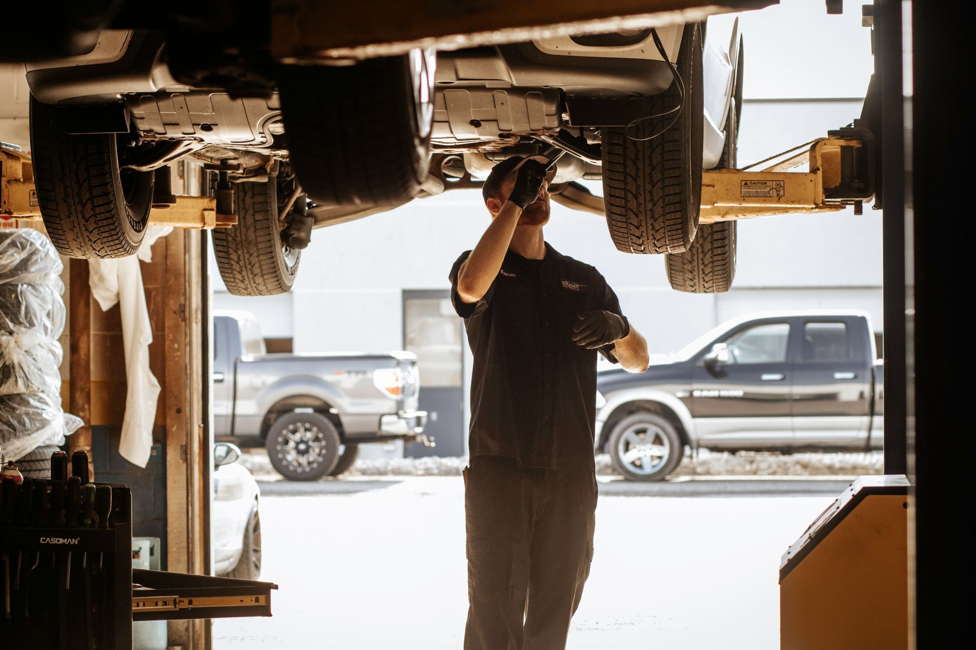 Mechanic working under a car on a lift in a shop, with two trucks in the background.