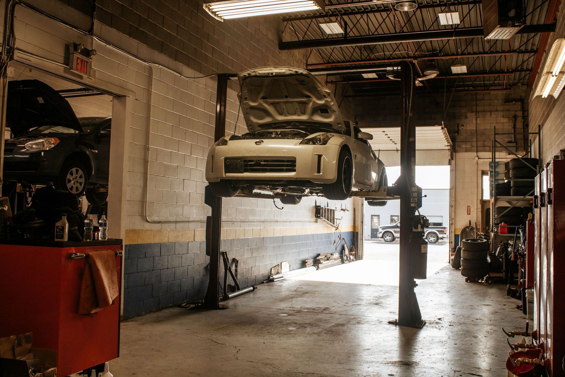 A white car on a lift inside a garage, hood open, with tools and another car visible.