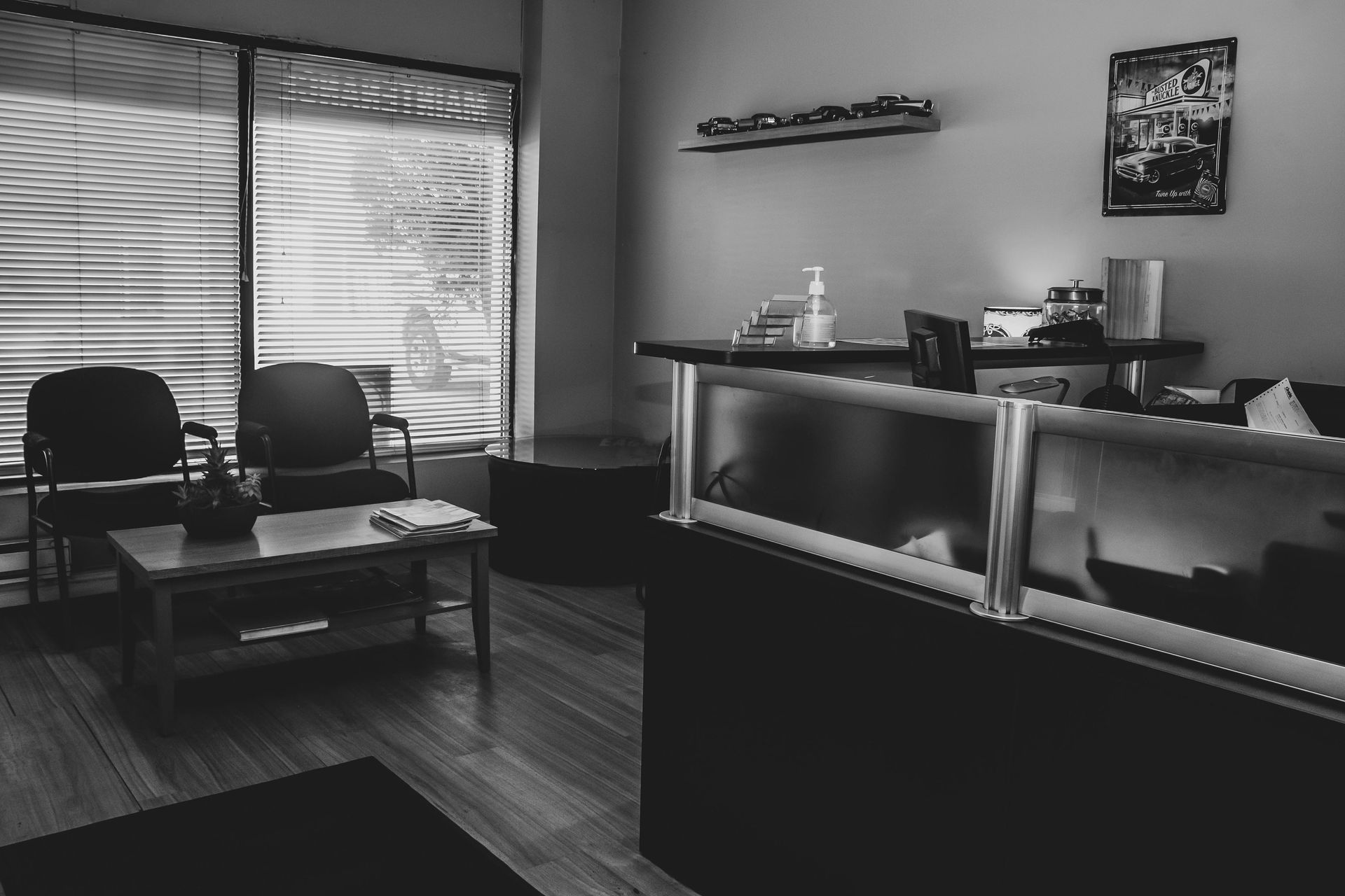 Black and white photo of a waiting room with chairs, a desk, and a window with blinds.