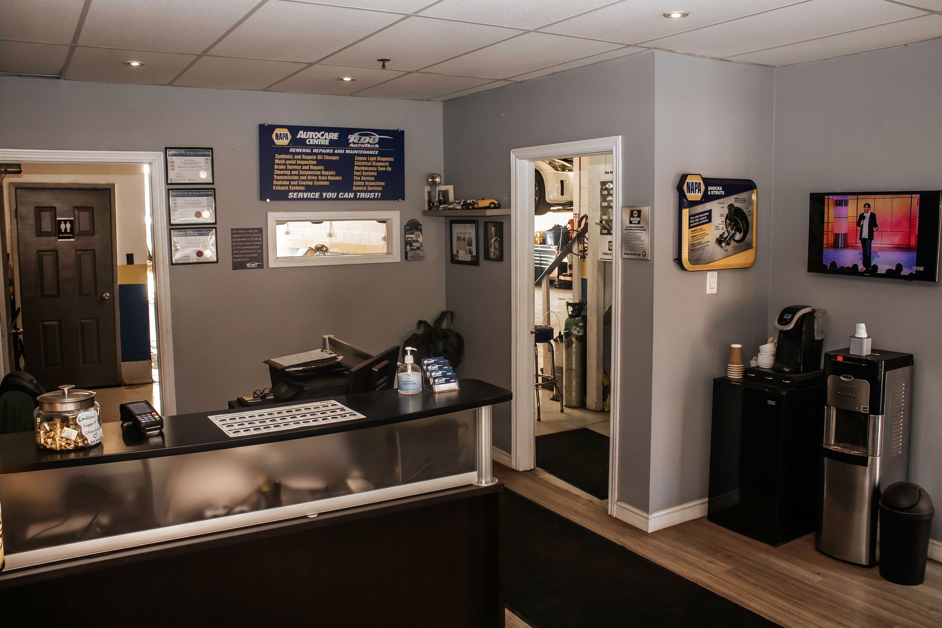 Reception area of an auto repair shop. Black countertop, gray walls, water cooler, TV, and waiting area.