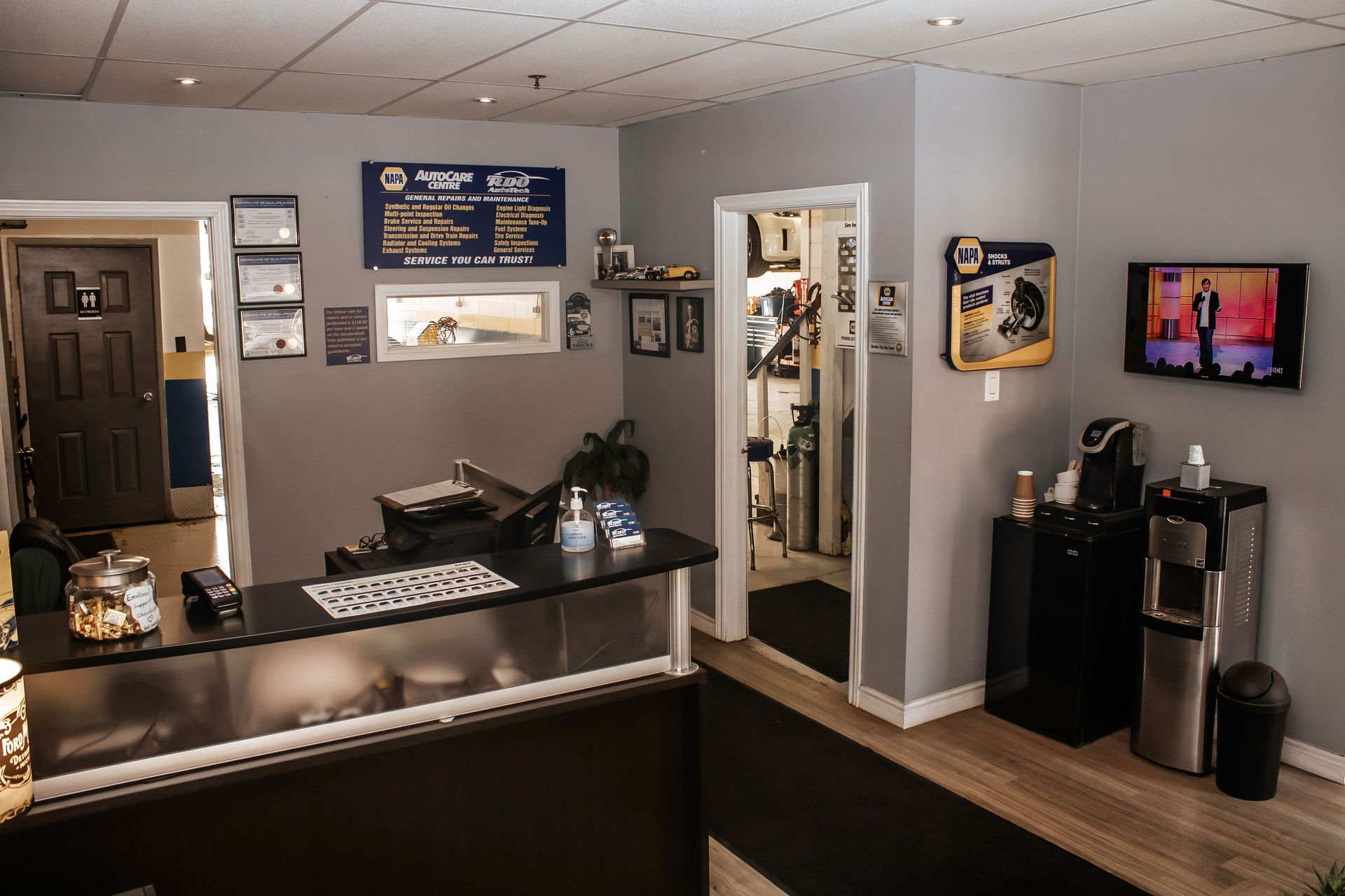 Reception area of an auto shop with a desk, water cooler, TV, and door leading outside.