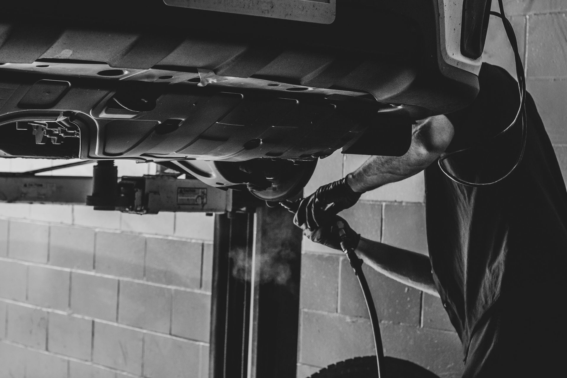 Mechanic working under a car on a lift, using a tool. Black and white, industrial setting.