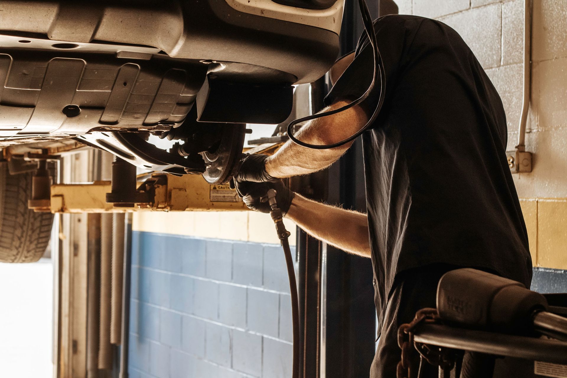 Mechanic working on a car lifted in a garage. Wearing gloves, black shirt, and using tools under vehicle.