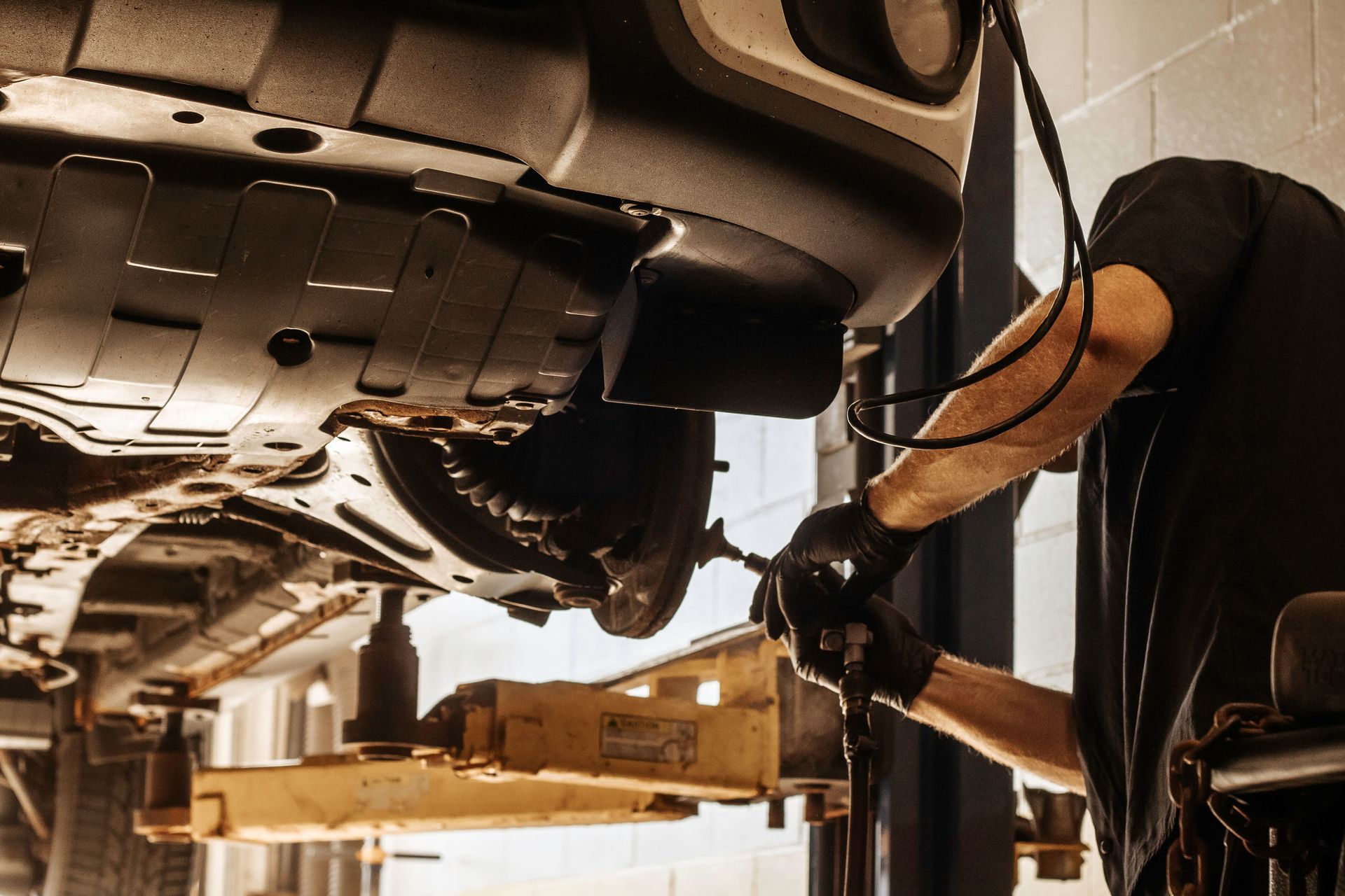Mechanic working on car, raised on a lift, in a garage; hands wearing gloves.