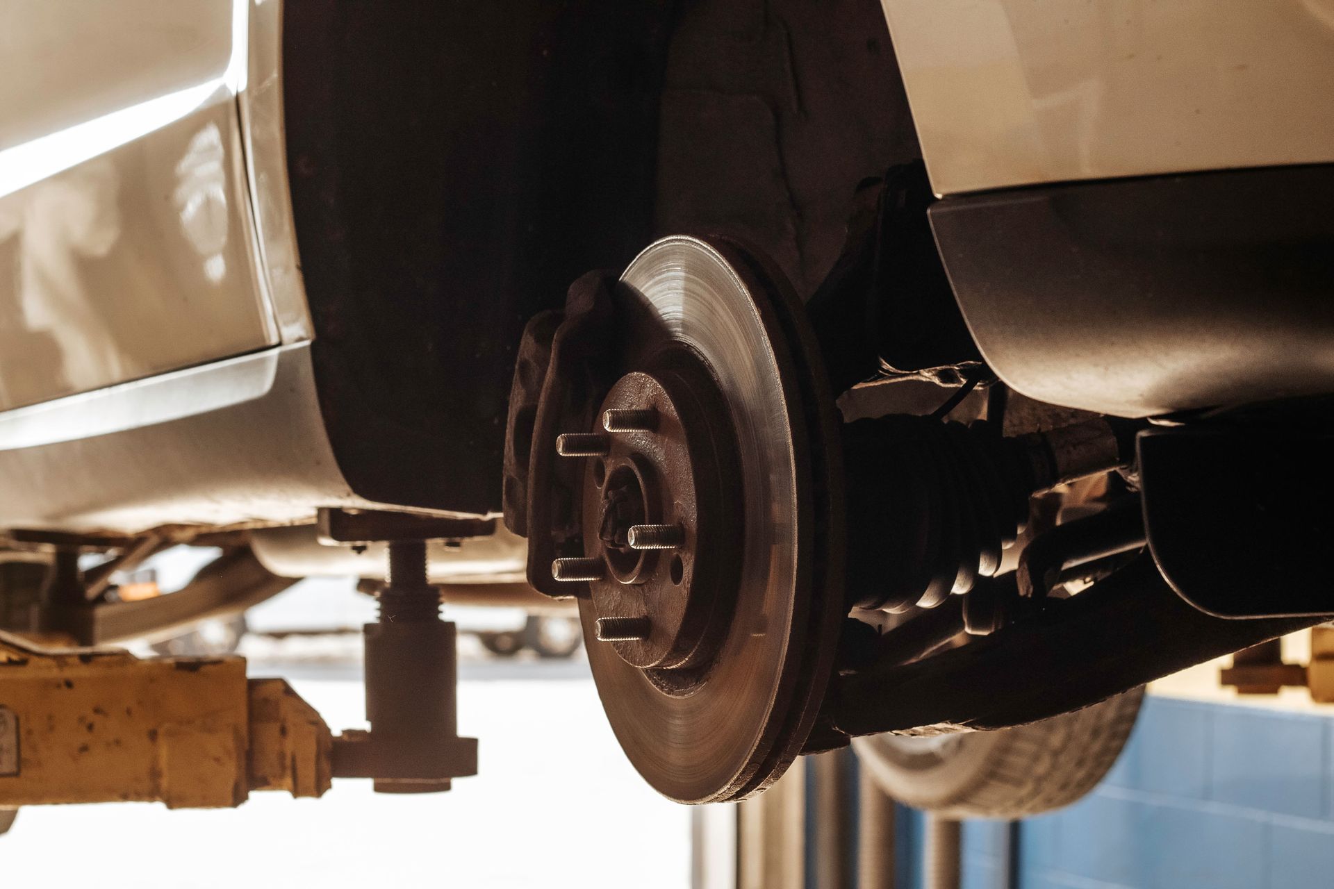 Car's front brake system on a lift. Silver car, brake rotor visible, yellow lift in a garage.