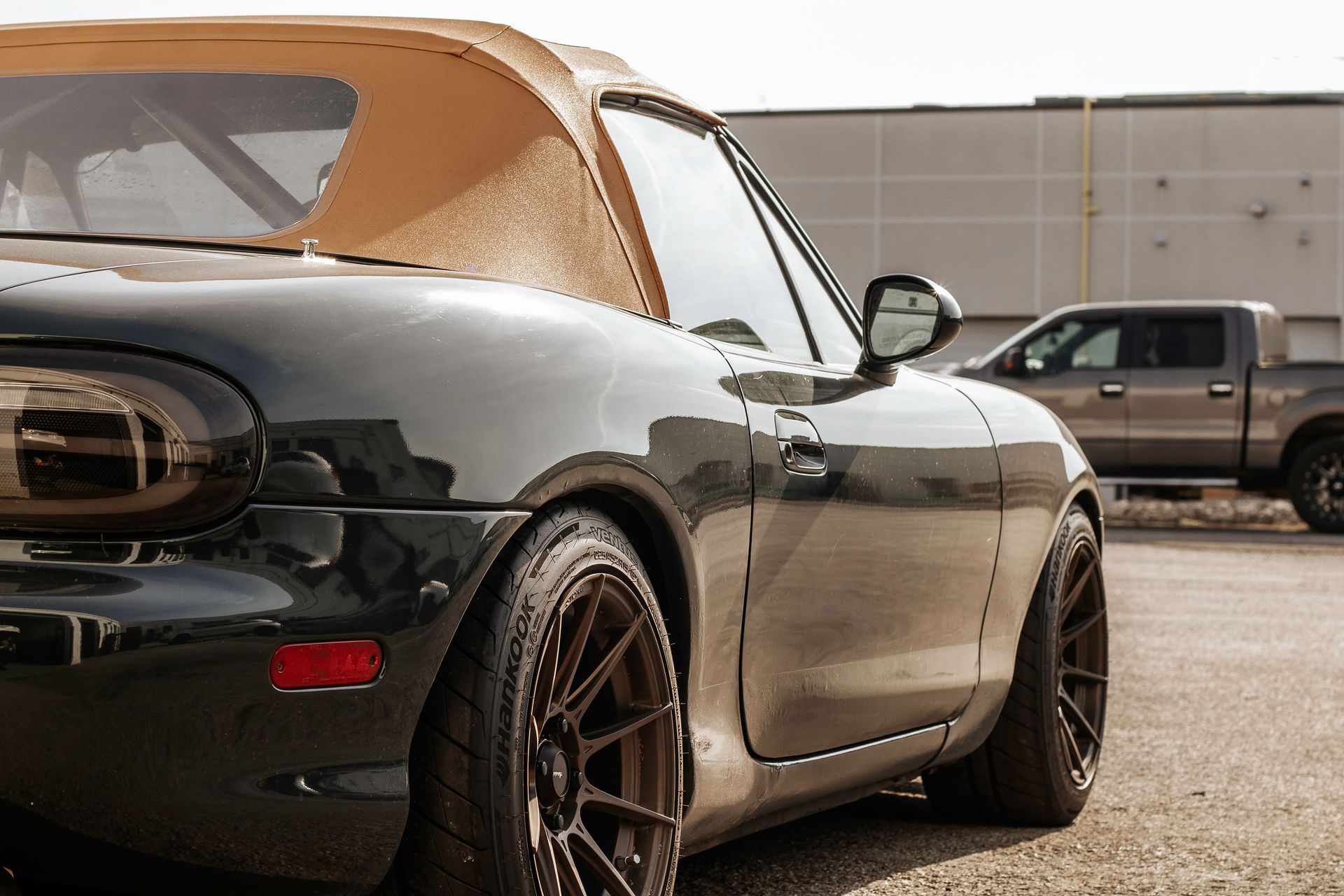 Black Mazda Miata convertible with brown wheels, top up, parked near a gray truck.
