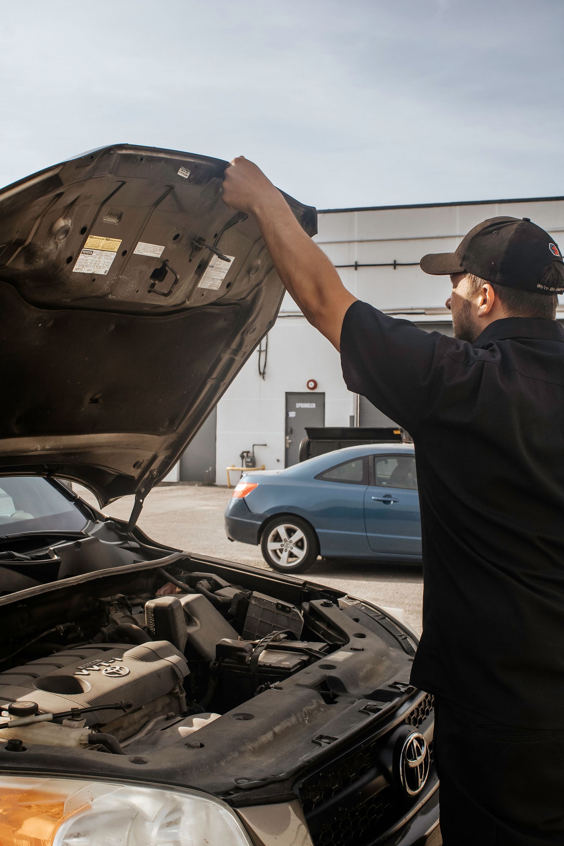 Mechanic lifting car hood, examining engine. Outdoors, blue car in background.