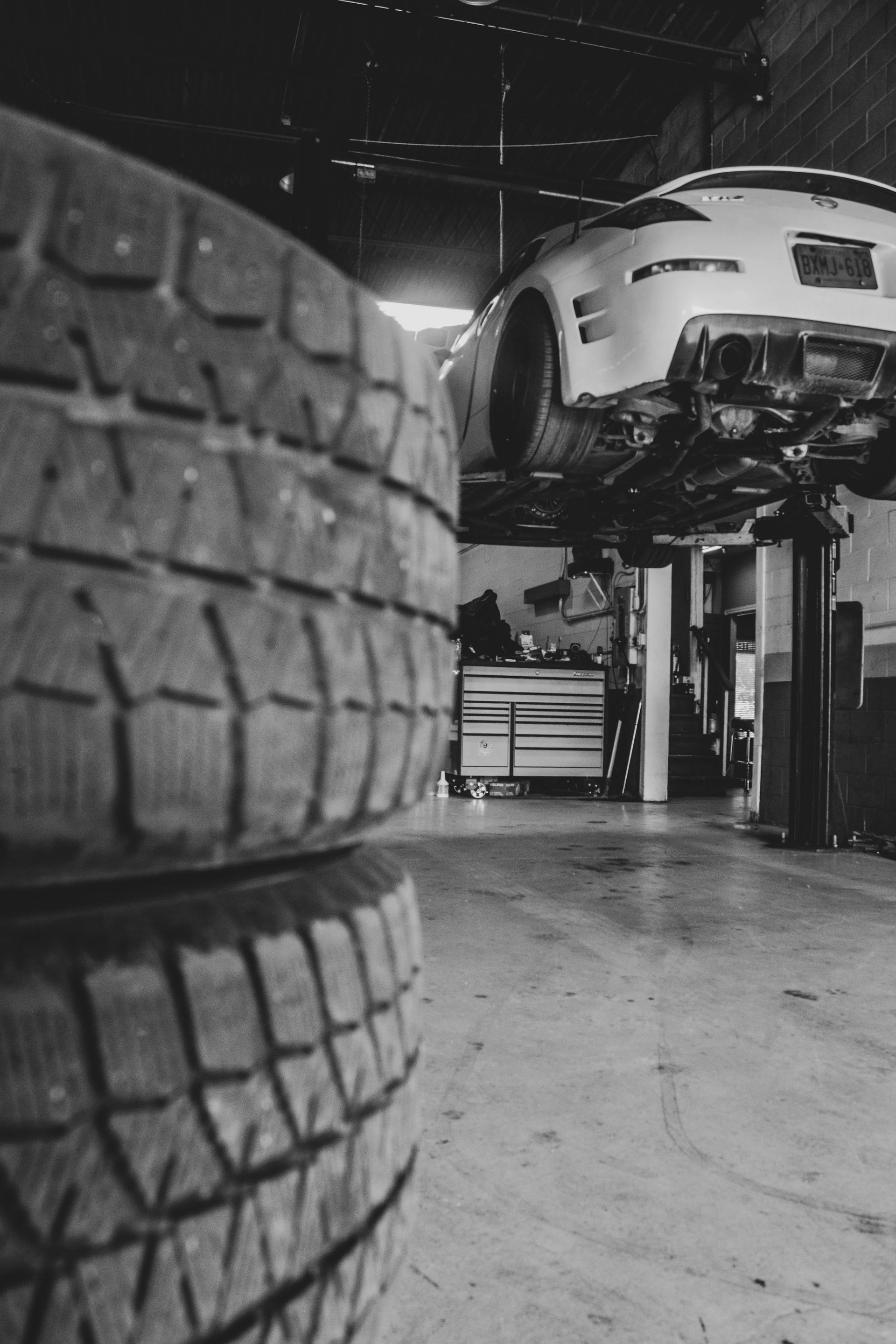 Tires stacked in front of a car raised on a lift in a garage; black and white.