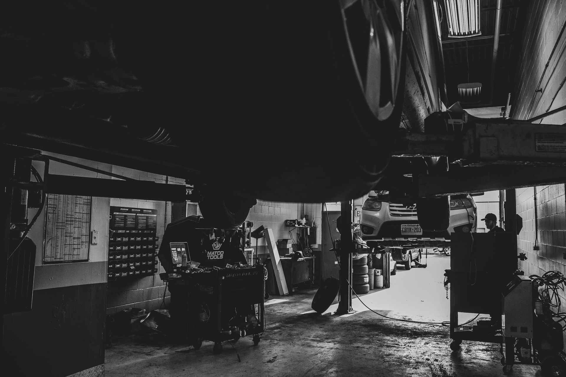 Car repair shop interior. A car is lifted, mechanics work below. Black and white.