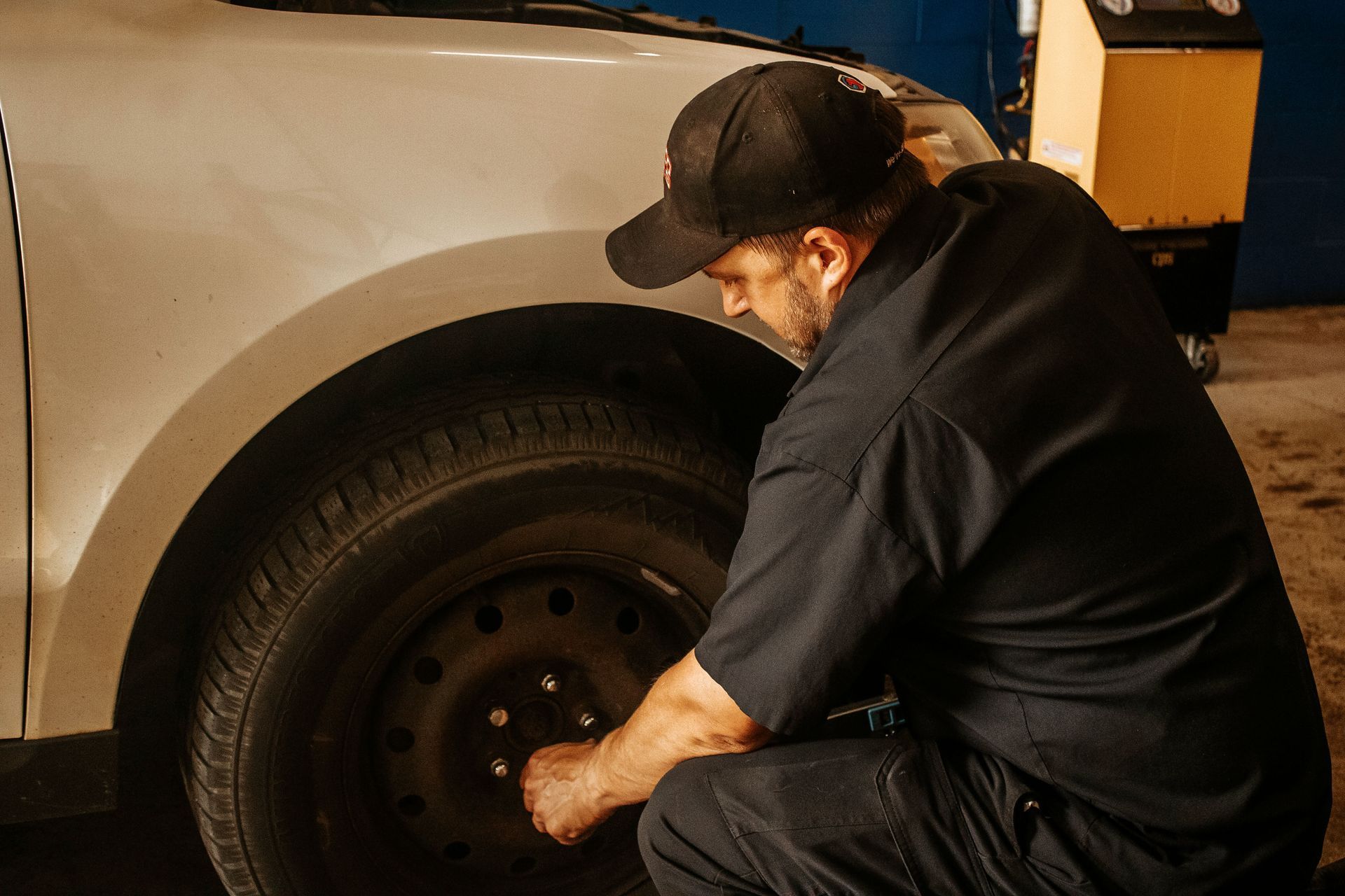 Mechanic in black shirt and hat changing a tire on a white car in a garage.