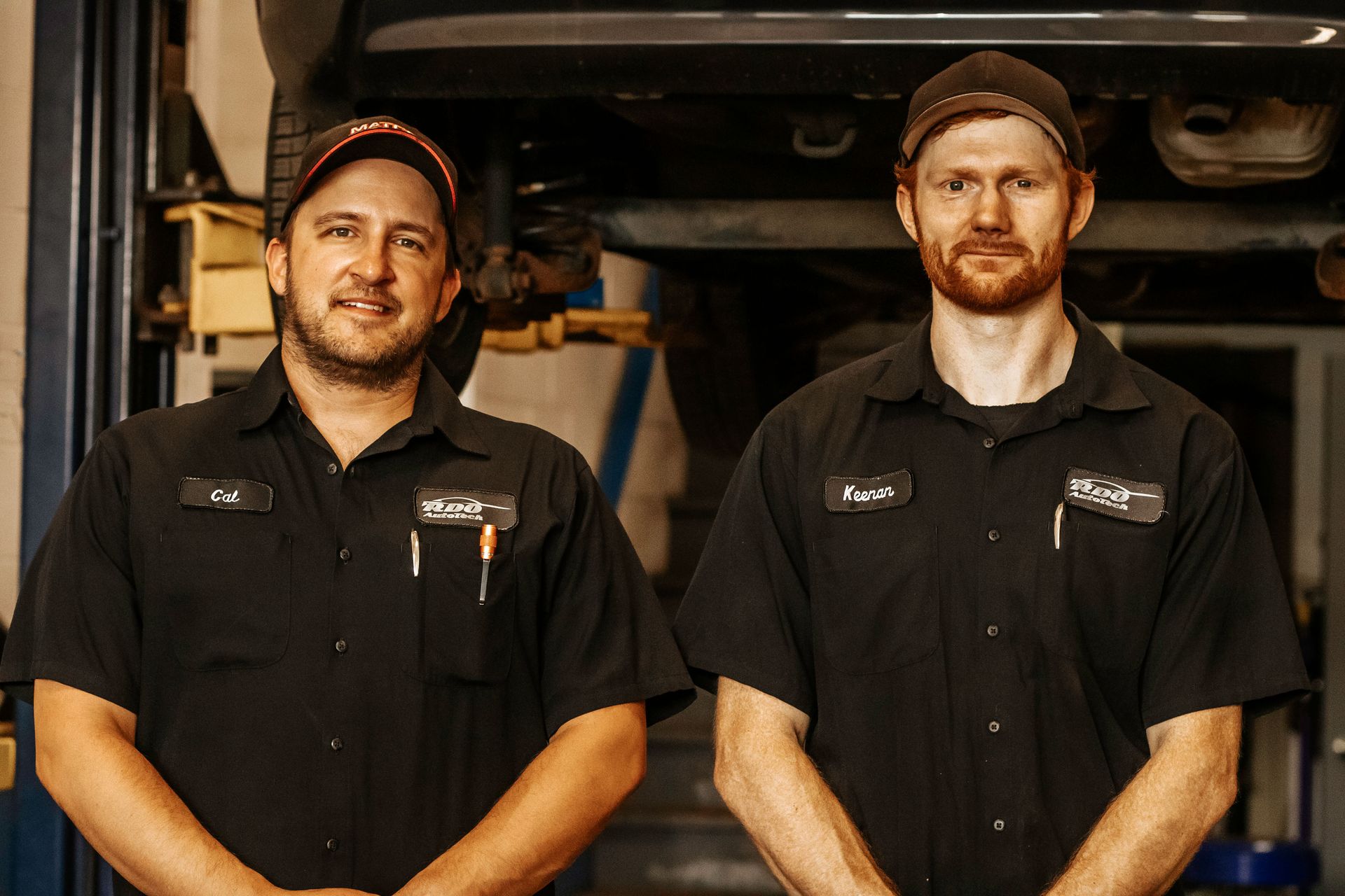 Two mechanics in black shirts, standing in a shop, look at the camera. One is smiling.