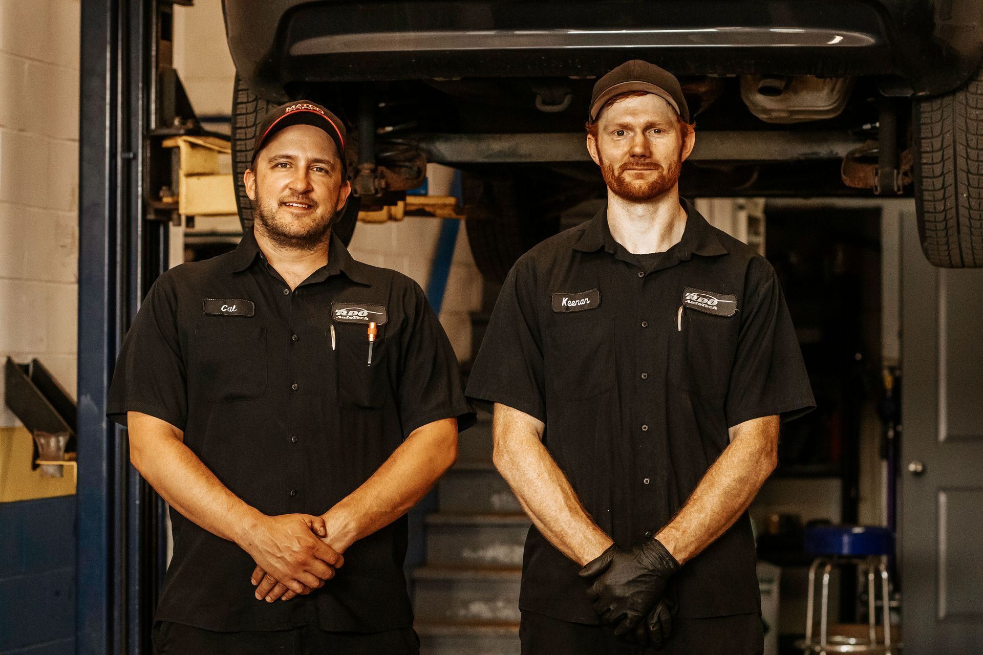 Two mechanics in black shirts pose in a garage under a lifted vehicle.