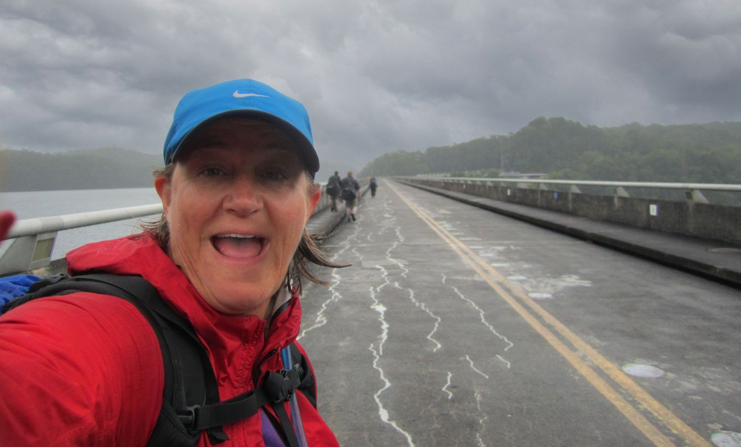 Woman with blue hat and red jacket smiles on a wet bridge, other hikers ahead. Overcast sky.