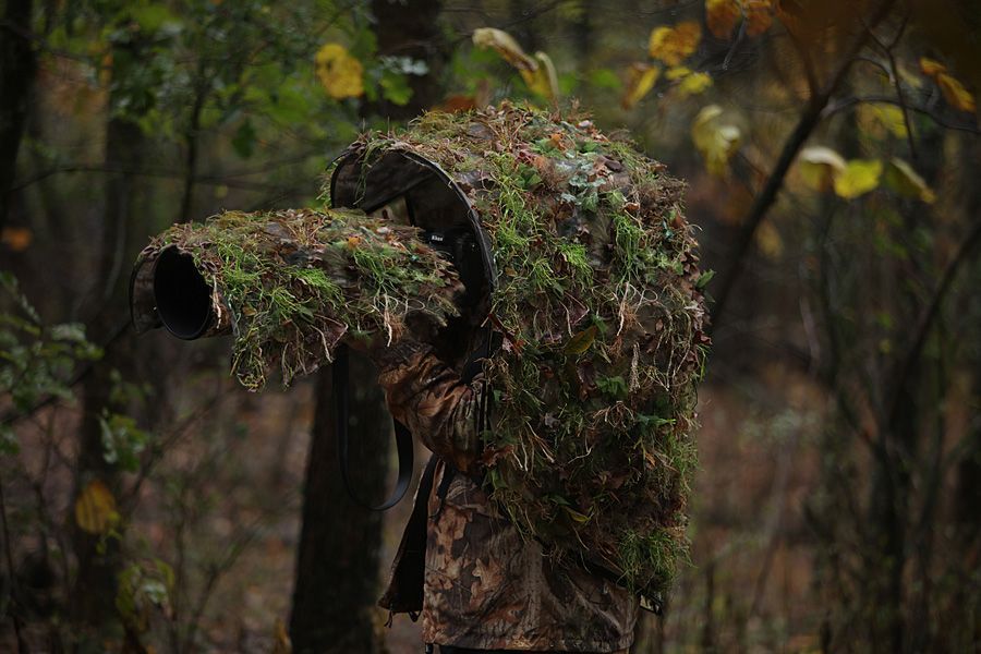 Photographer in GOShelter hands-free canopy with Ghillie 3D camouflage skin holding a large camera with the GOShelter Telescopic Sleeve with Ghillie 3D camouflage attached, set in a forest.