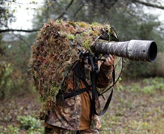 Photographer in GOShelter hands-free canopy with Ghillie 3D camouflage skin holding a large camera, set in a forest.